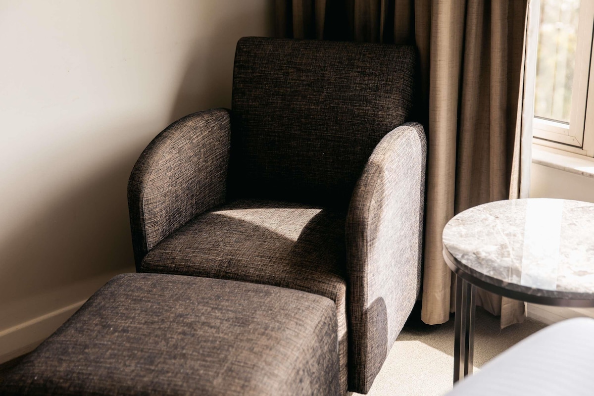A room with a dark grey upholstered armchair, a small round table, and a window with curtains.
