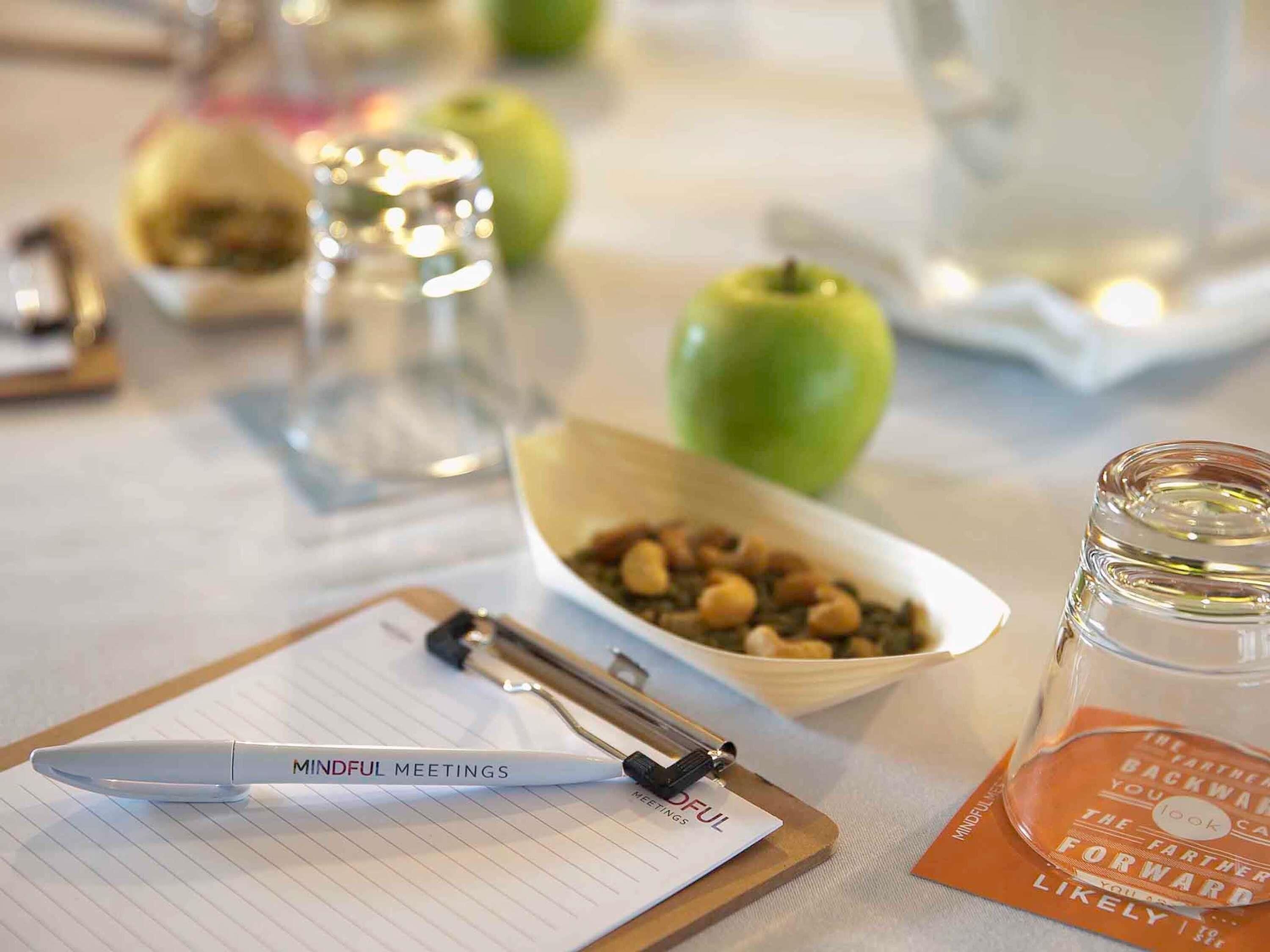 Una pissarra amb la inscripció 'Mindful Meetings', una ploma, un bol de fruits secs, una poma verda i un bocal de vidre amb una etiqueta.