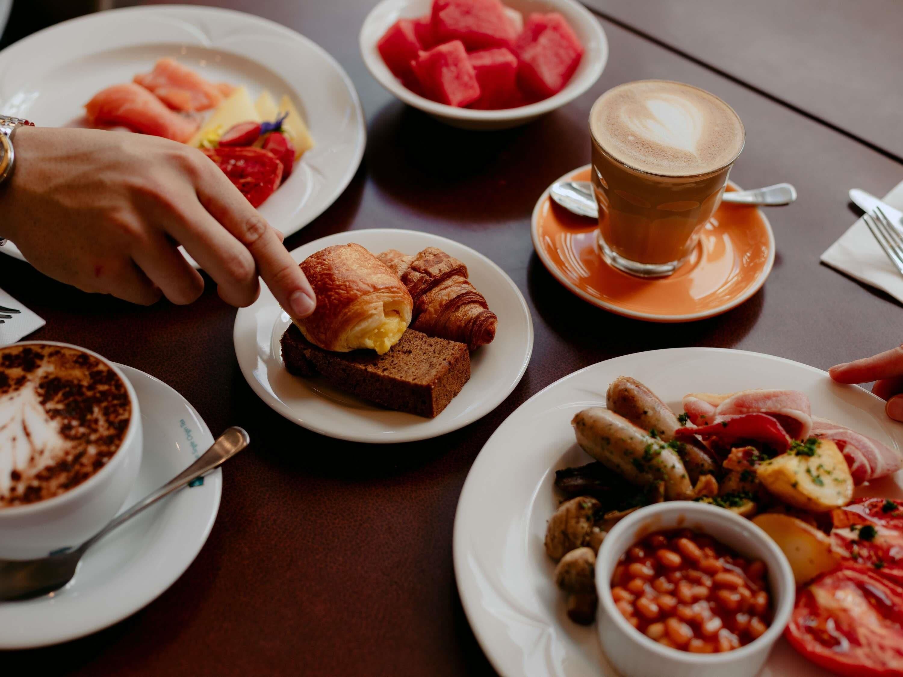 Et bord med forskellige morgenmadsretter, blandt andet croissanter, en kop kaffe, vandmelon og en tallerken med bønner og grøntsager.