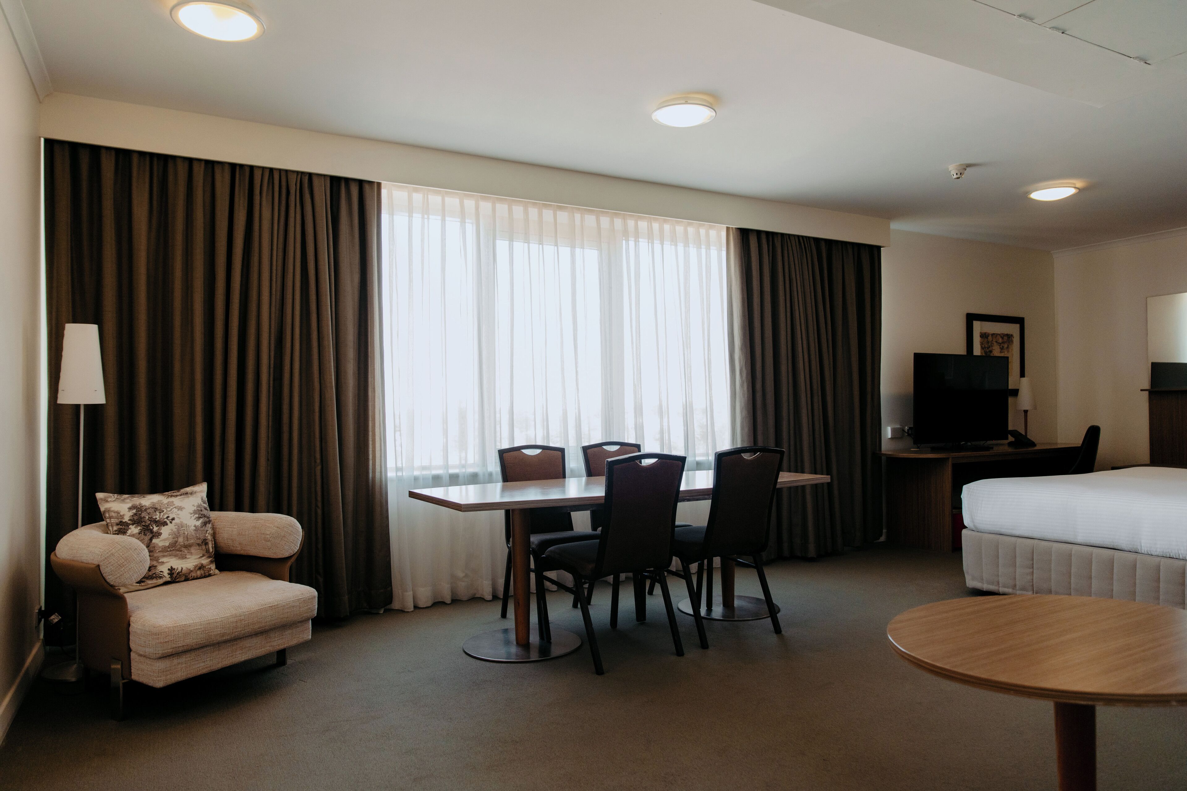 A hotel room with a bed, a desk with a television, a dining table with chairs, and a sofa.