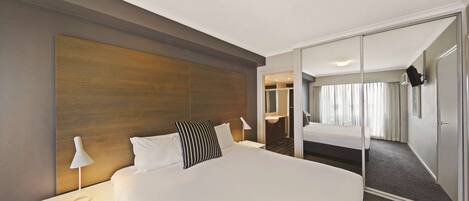 Une chambre d’hôtel avec un grand lit, une table de chevet avec une lampe, une tête de lit en bois, un miroir et une armoire.