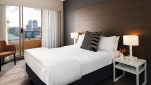 Habitación de hotel con una cama grande, dos lámparas de noche, una mesita de noche, una silla y vistas a la ciudad a través de amplios ventanales.