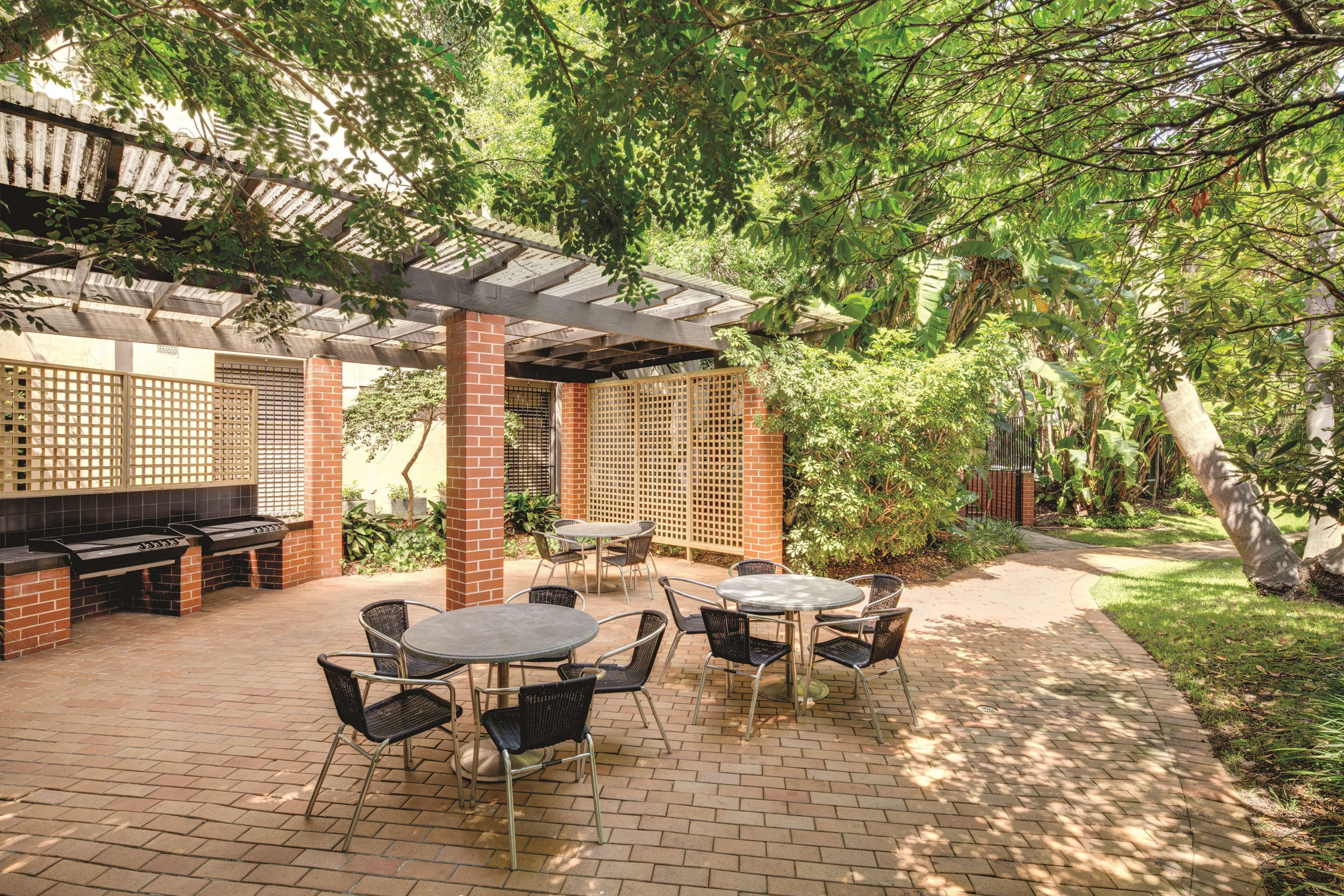 Een terras buiten met bakstenen vloer, metalen tafels en stoelen, een barbecue en een pergola met een rasterwerk.