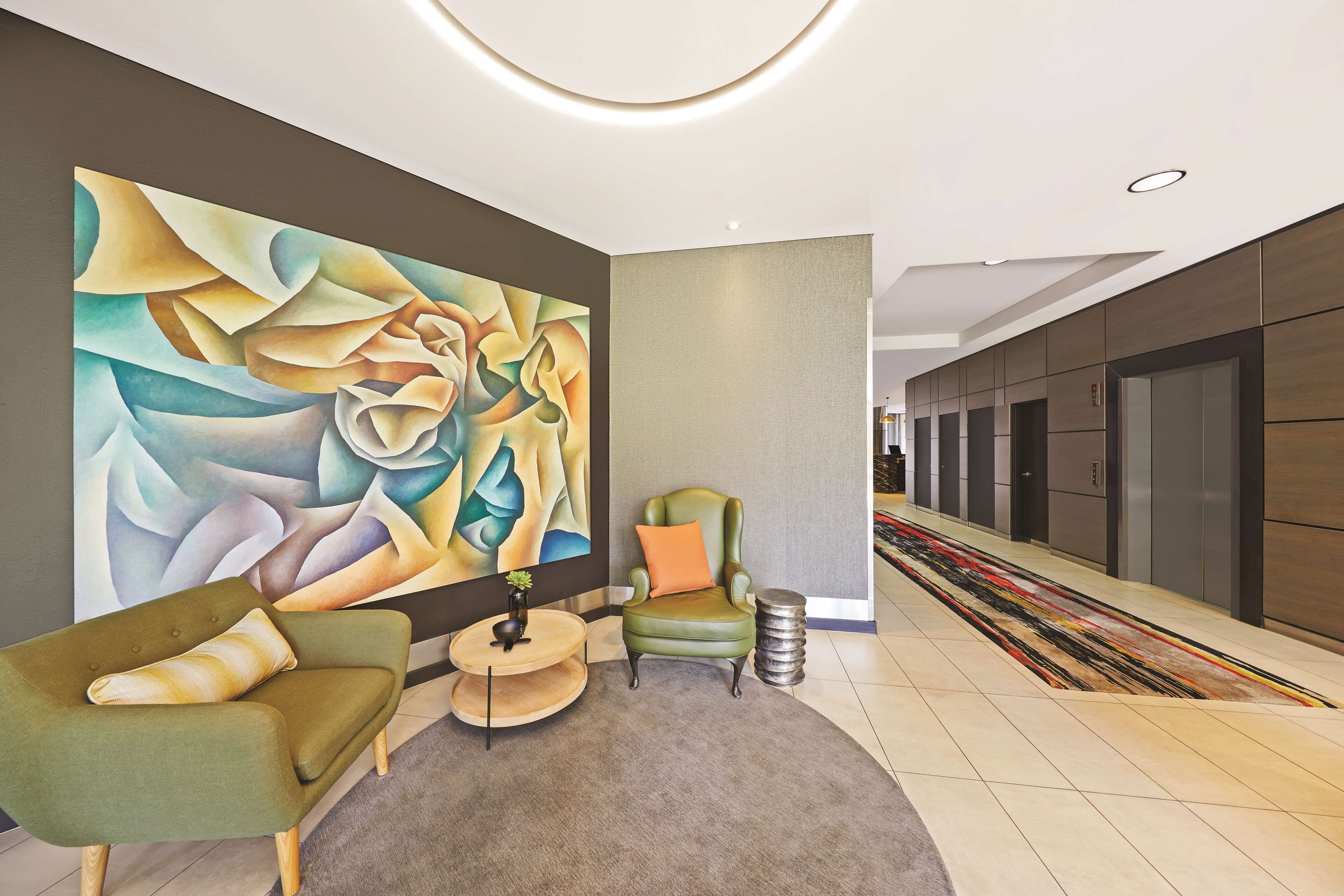A lobby with a green armchair, a small table with a plant, and a colorful abstract painting on the wall.