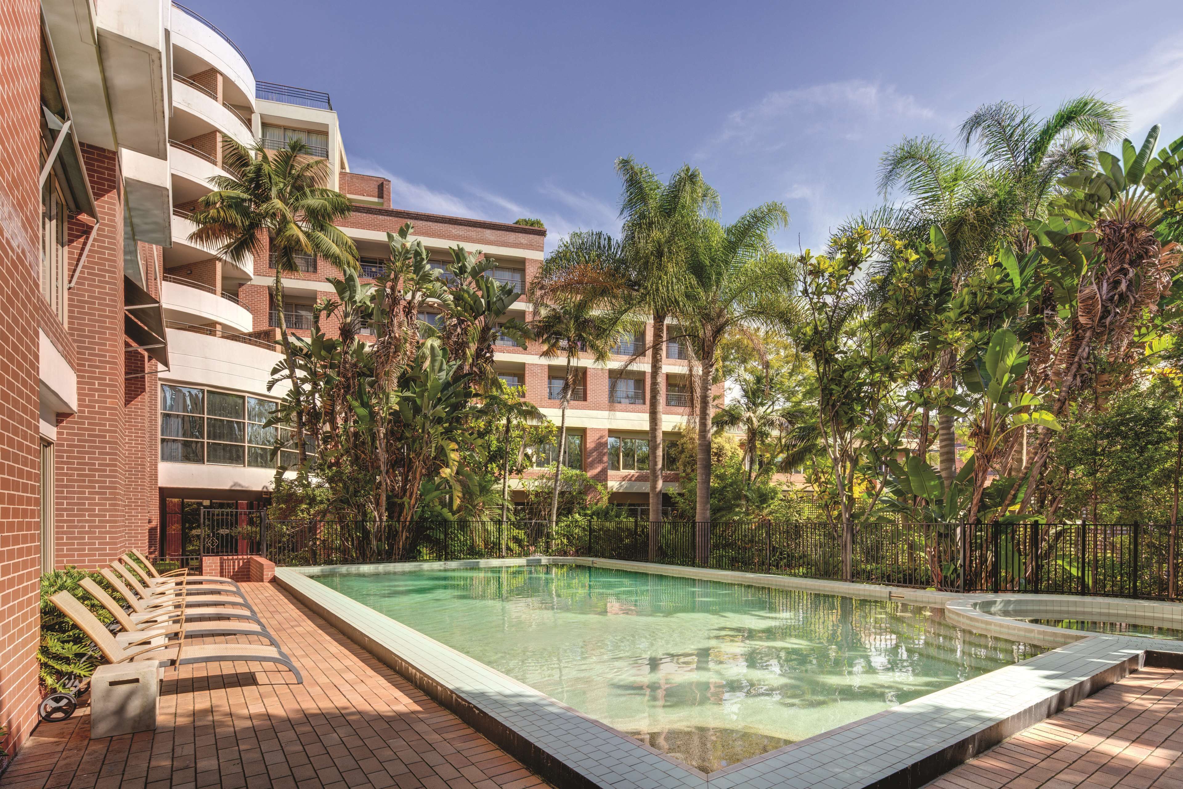 A pool area with lounge chairs, surrounded by palm trees and a multi-story building.