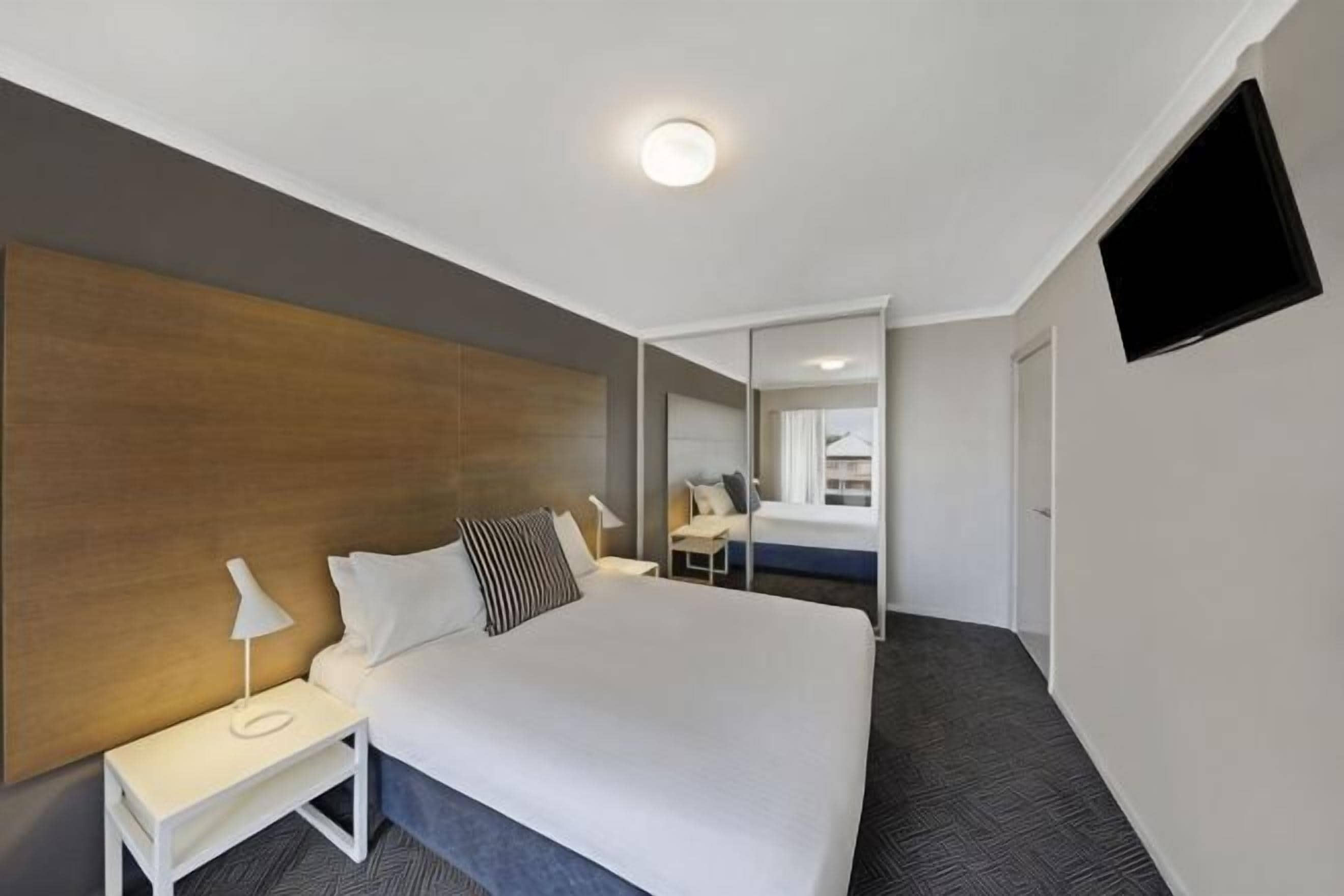 Een hotelkamer met een groot bed, nachtkastje met een lamp, een flatscreen televisie aan de muur en een spiegel die de kamer weerspiegelt.