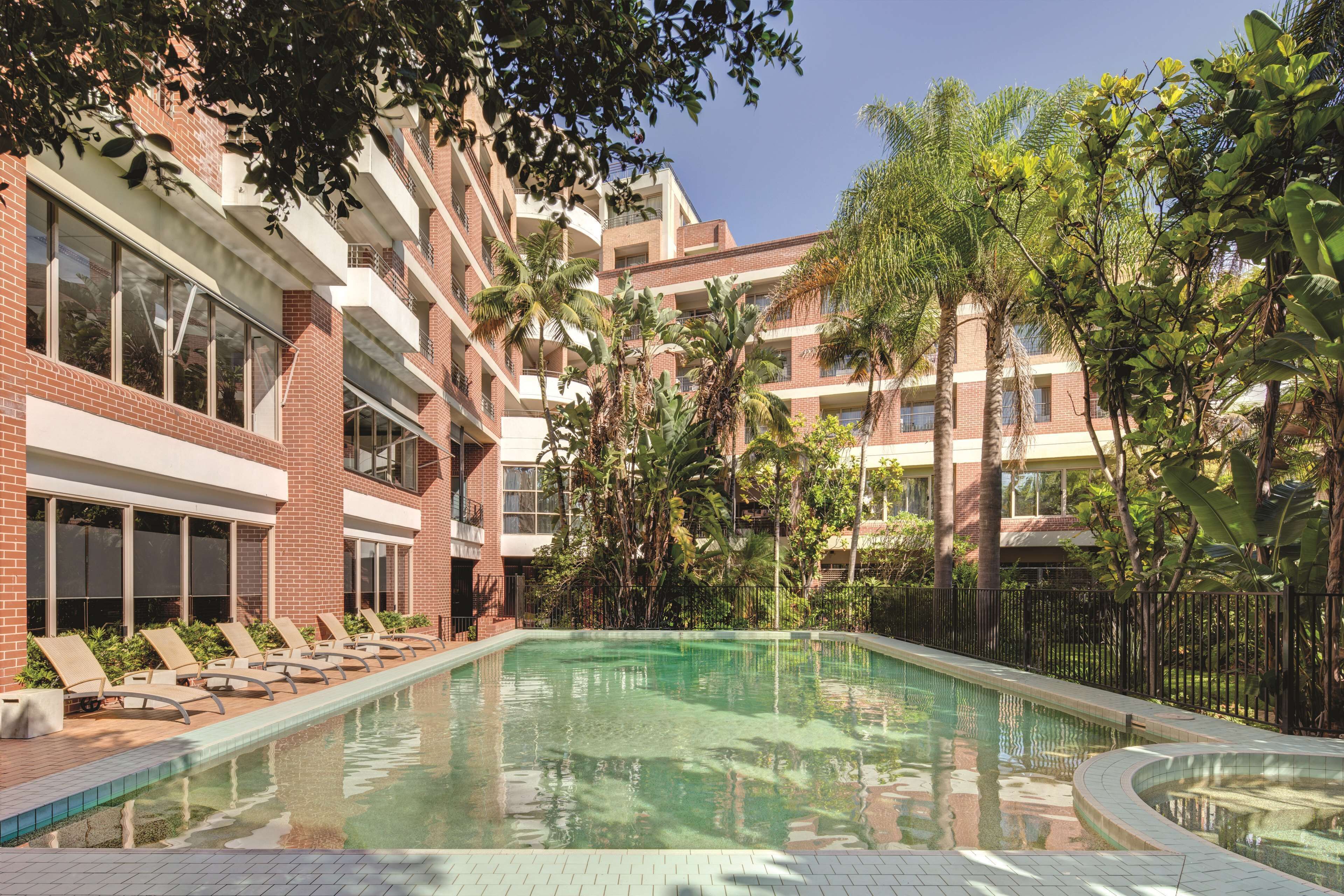A swimming pool surrounded by lounge chairs and palm trees, with a multi-story building in the background.