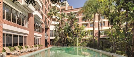 A swimming pool surrounded by lounge chairs and palm trees, with a multi-story building in the background.