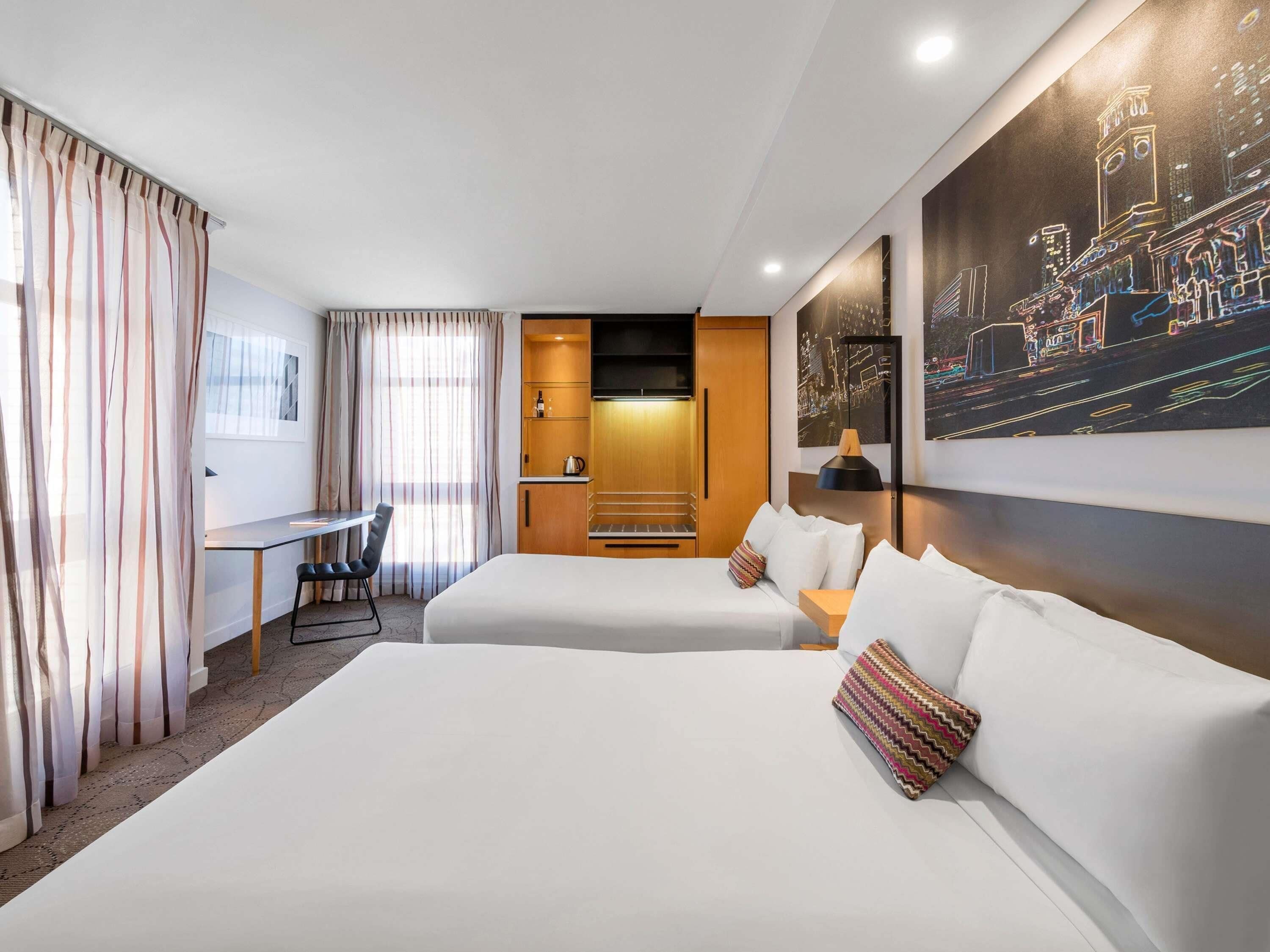 Habitación de hotel con dos camas, un escritorio, una silla, una kitchenette pequeña y un cuadro con una vista urbana.