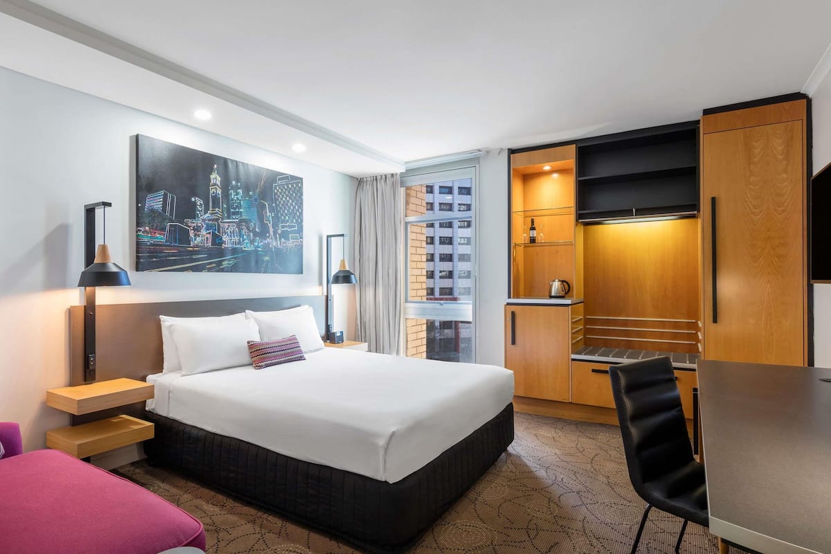 A hotel room with a bed, a desk, a chair, a TV, a painting, and a kitchenette.