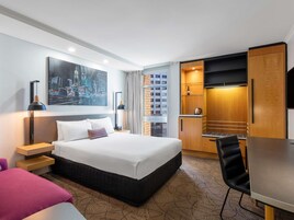 Hotellihuone, jossa on sänky, työpöytä, tuoli, televisio, taulu ja pieni keittiönurkkaus.