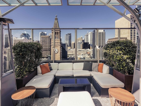 Un lounge en la azotea con vistas a la ciudad, un sofá modular grande y dos mesas redondas pequeñas.