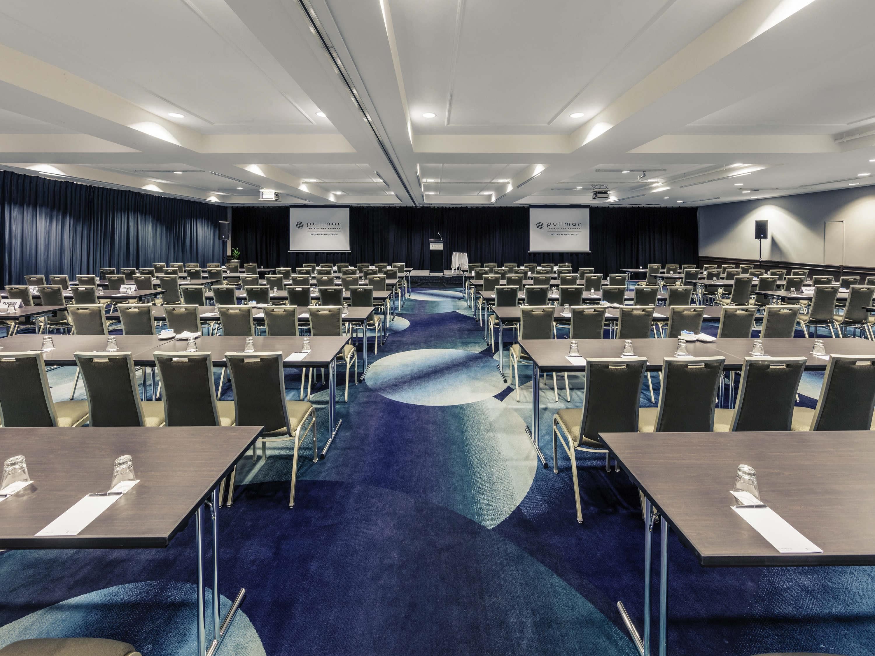 Un amplio salón de conferencias con filas de mesas y sillas, un escenario y una pantalla que muestra el nombre del hotel.