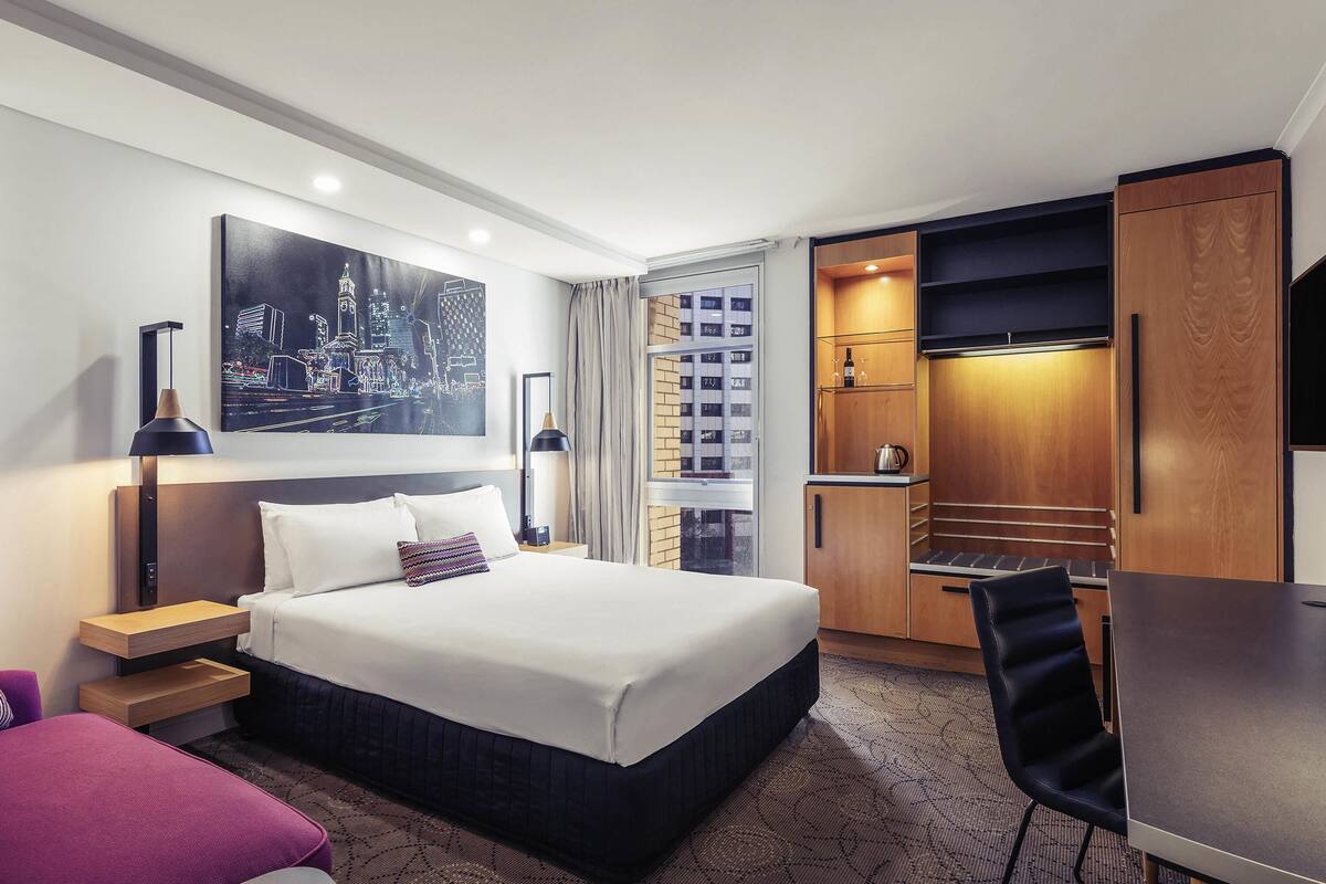 Habitación de hotel con cama, mesitas de noche, escritorio, silla, cuadro, lámpara, minibar y armario.