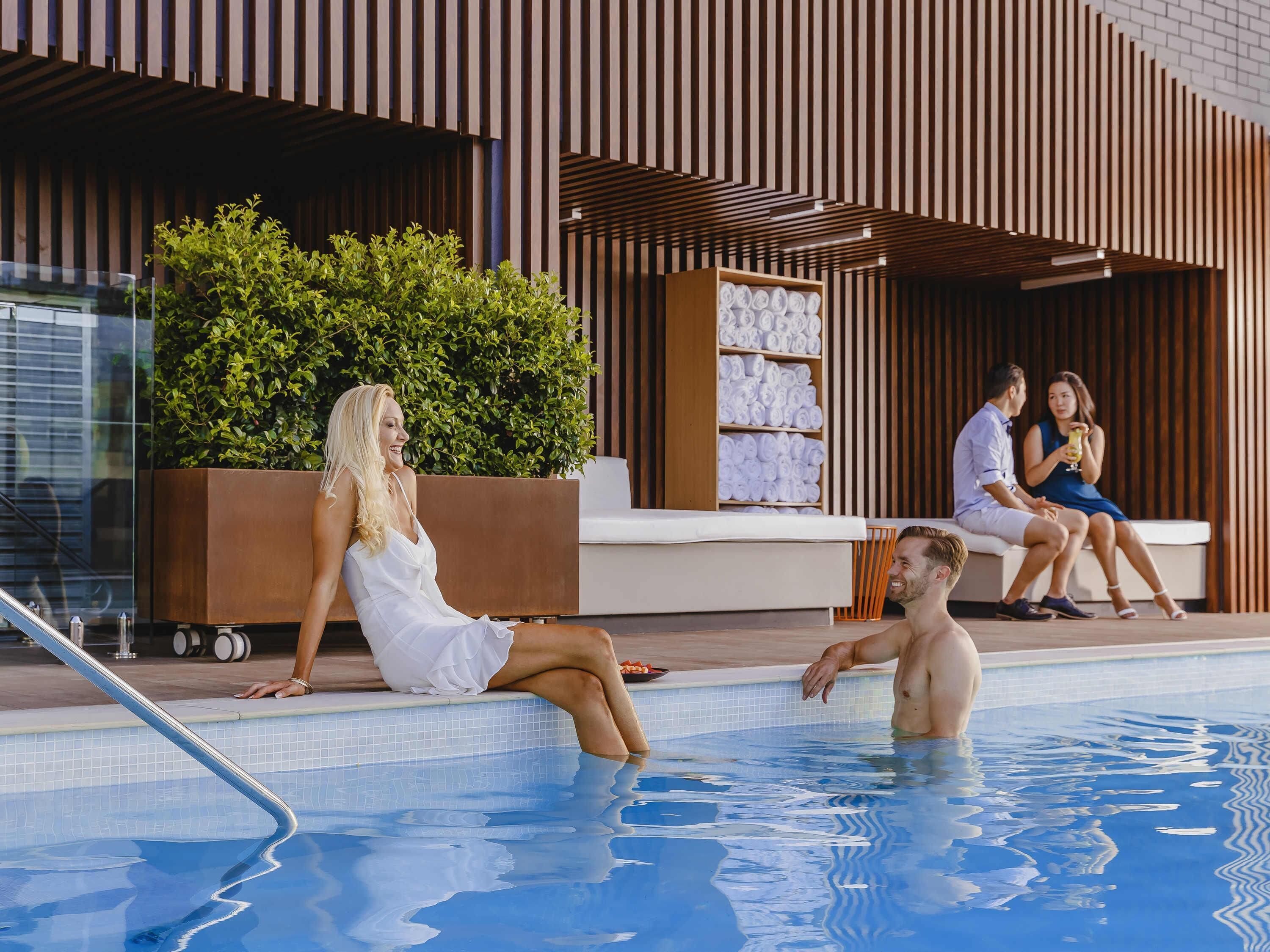 A poolside area with a modern building, a seating area, and a person sitting by the pool.