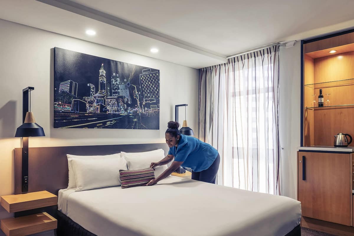 A hotel room with a neatly made bed, a cityscape painting on the wall, a wooden cabinet with a kettle, and a nightstand with a lamp.