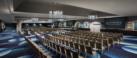 A conference room with rows of chairs, a stage, and a large screen displaying the hotel name.