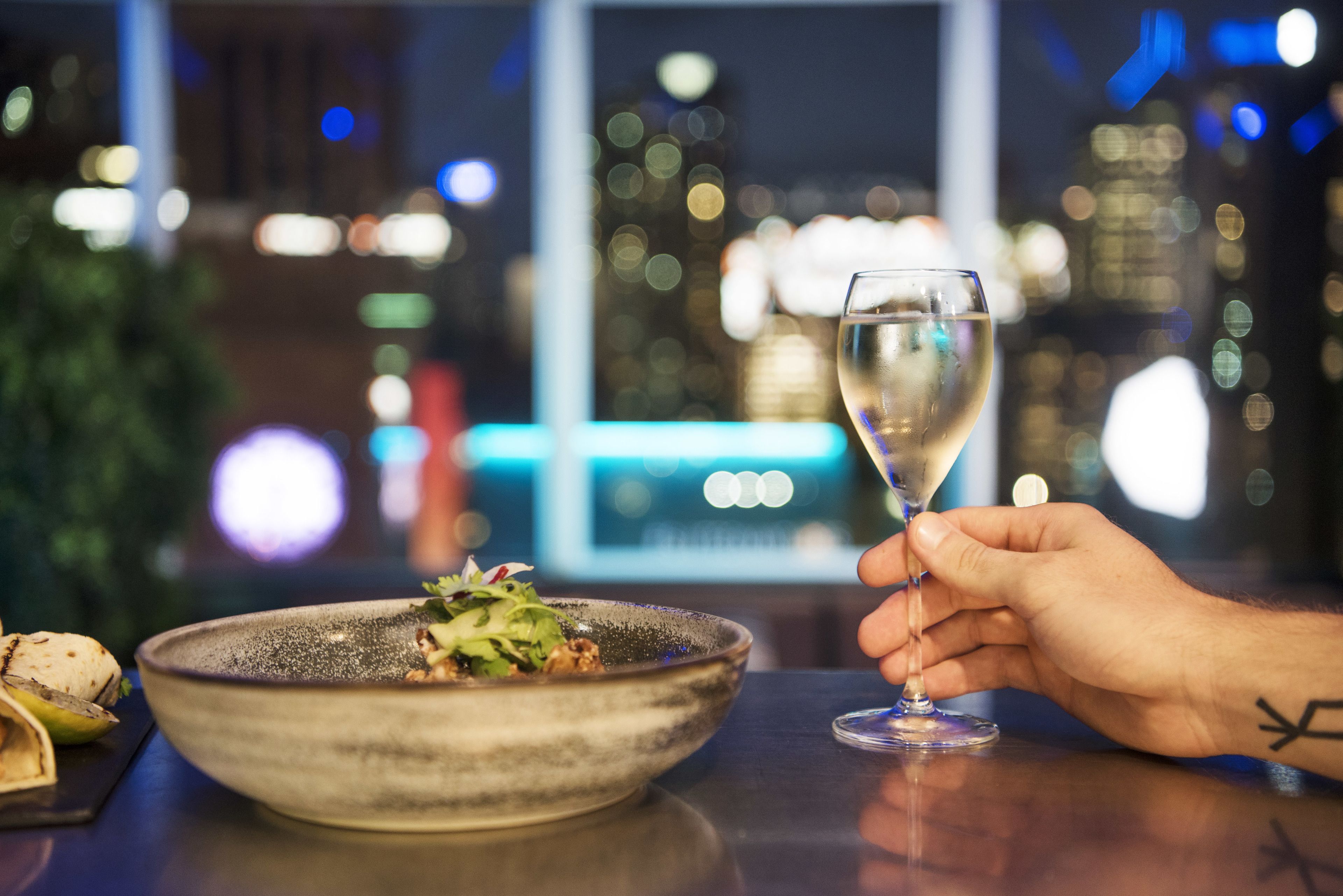 Ein Esstisch mit einer Salatschüssel und einem Glas Weißwein, vor verschwommener Stadtkulisse.