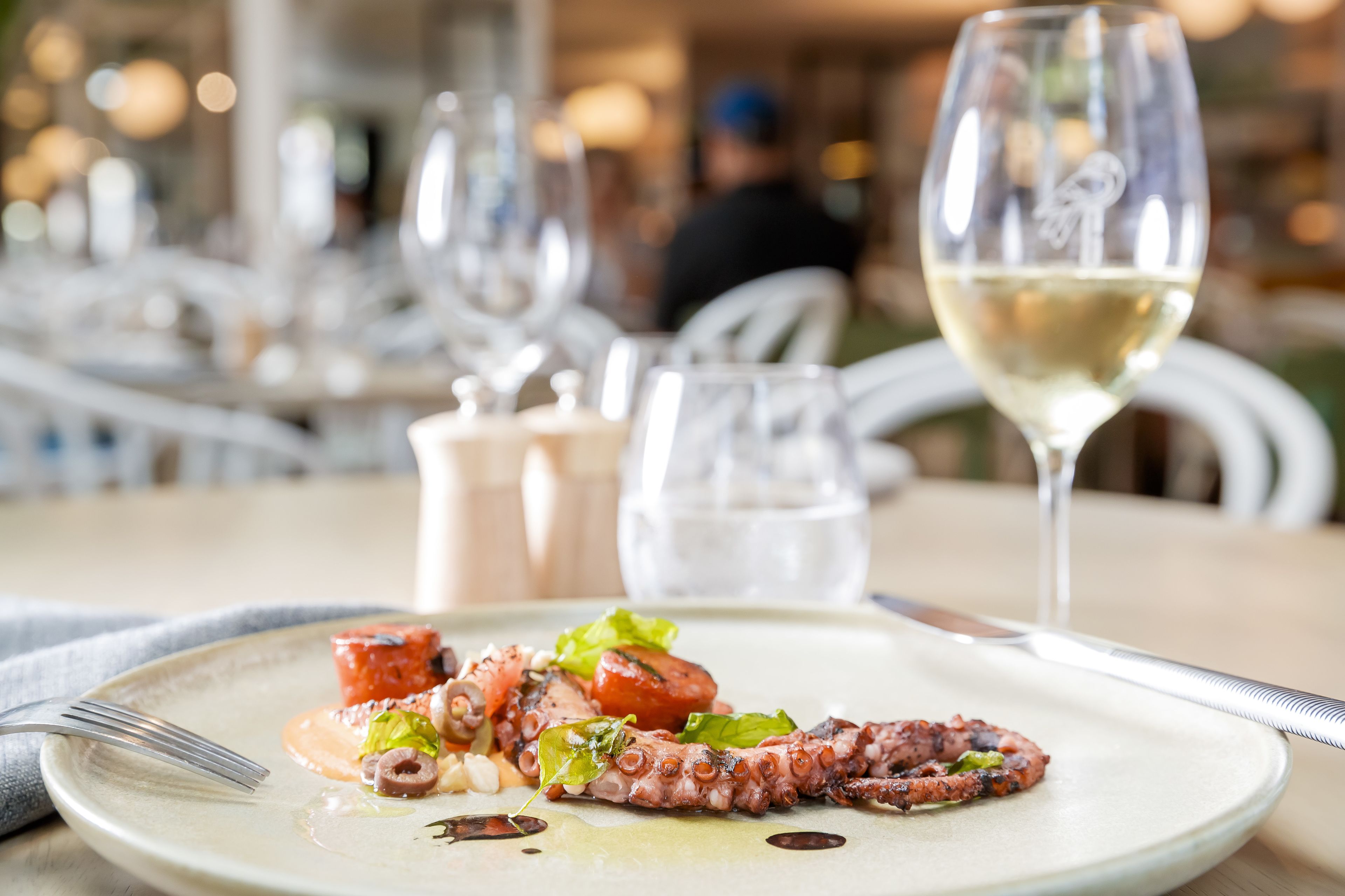 A dining table with a plate of octopus and vegetables, a glass of white wine, and a fork.