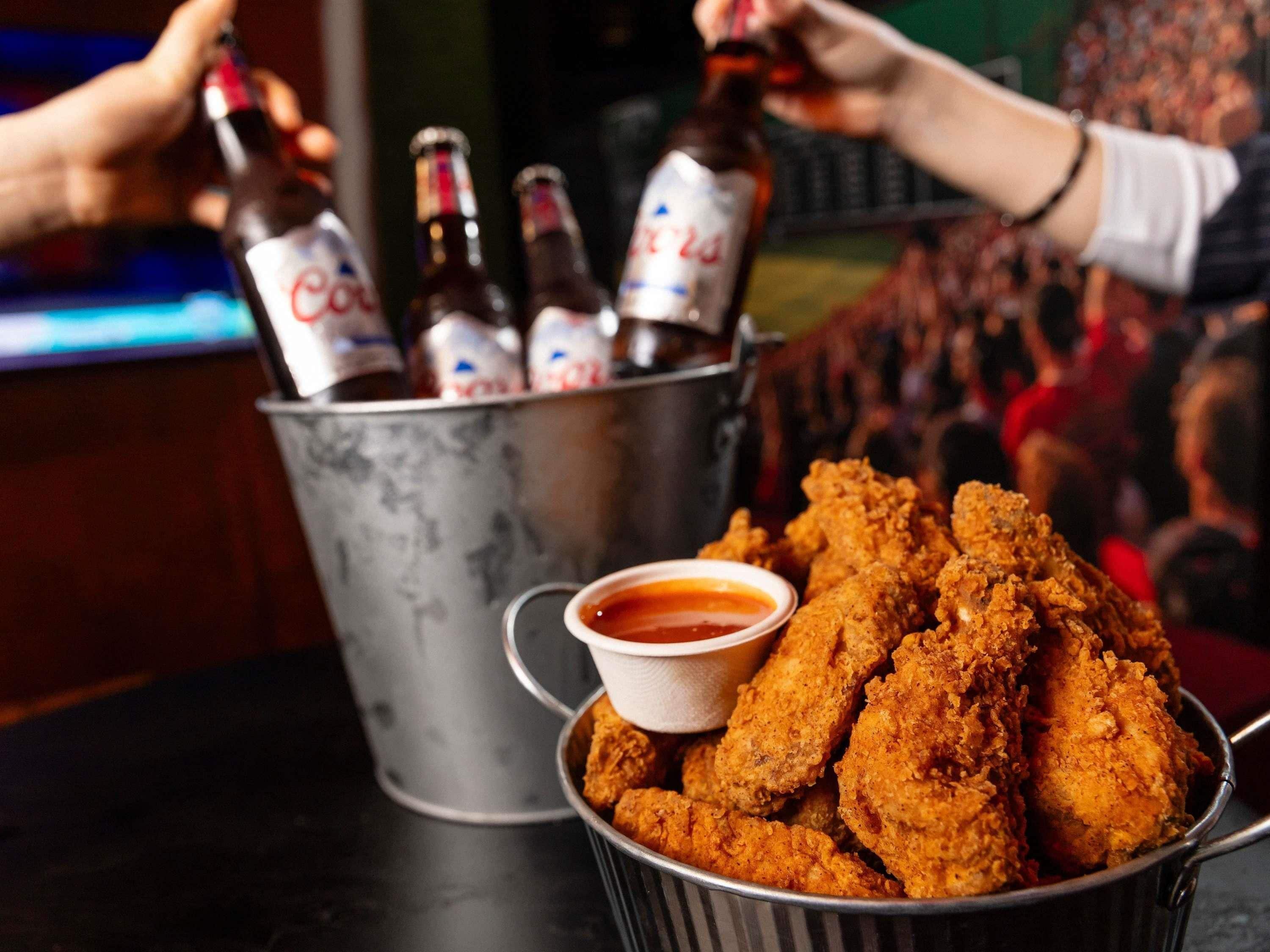 A bucket of fried chicken with a small bowl of sauce, and several bottles in the background.