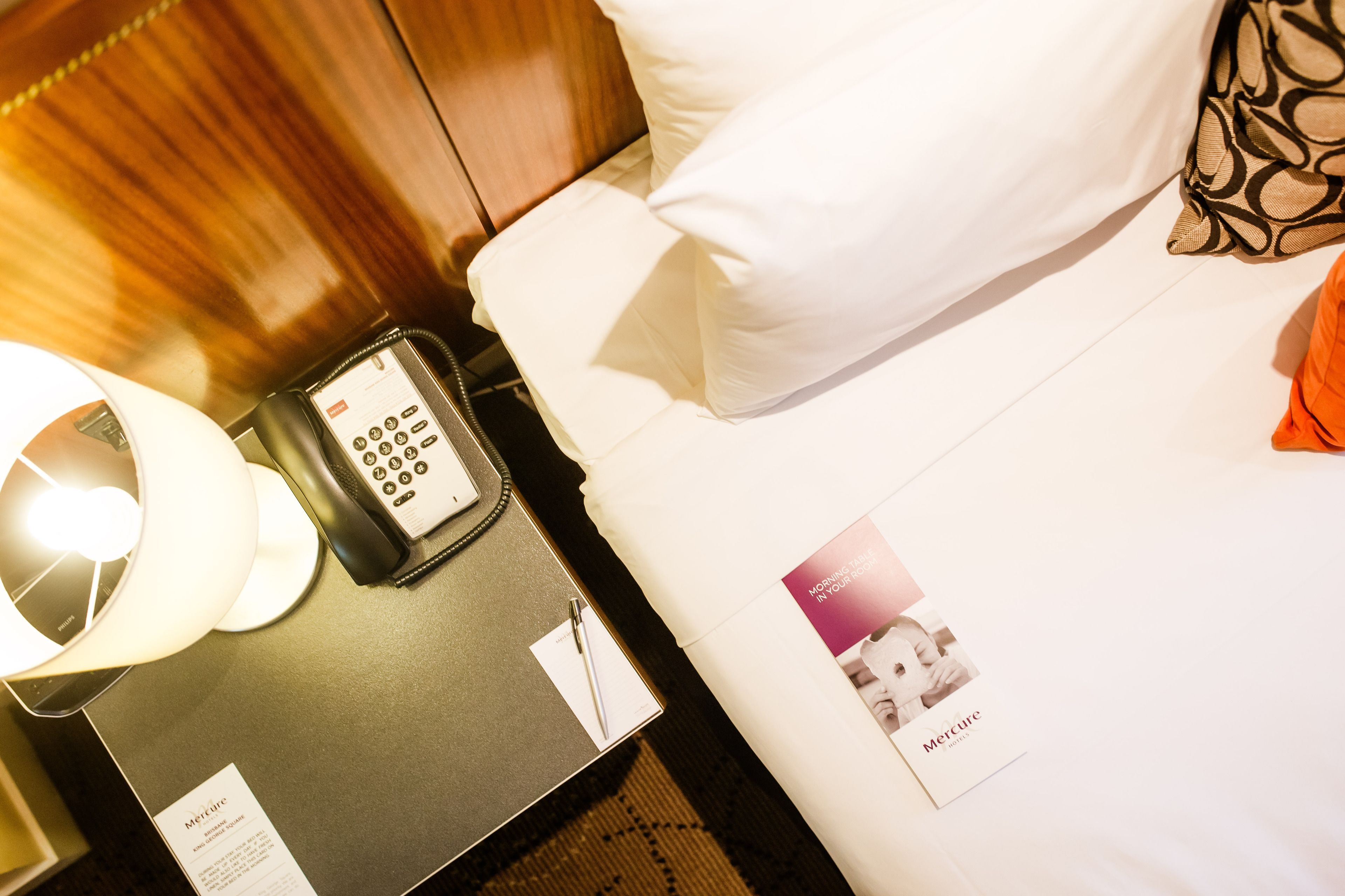 A hotel room with a bedside table, a telephone, a lamp, a pillow, and a brochure.