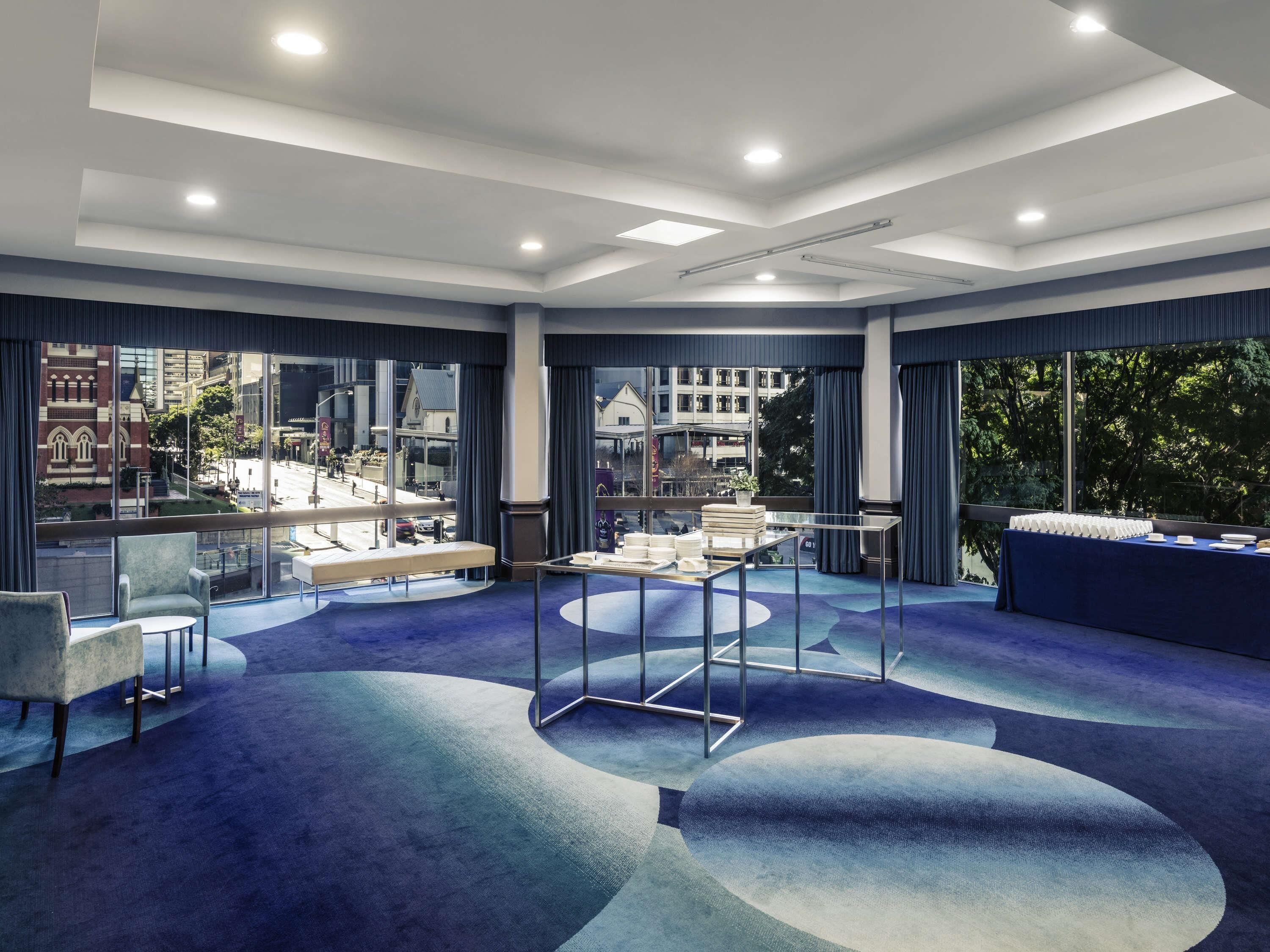 A modern hotel room with large windows, a blue carpet, and a seating area with a table.