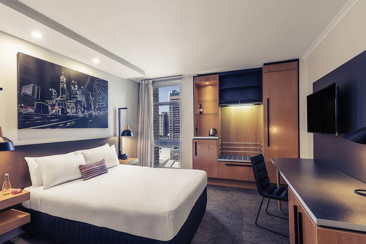 A hotel room with a bed, a desk, a TV, a painting, and a cityscape view.