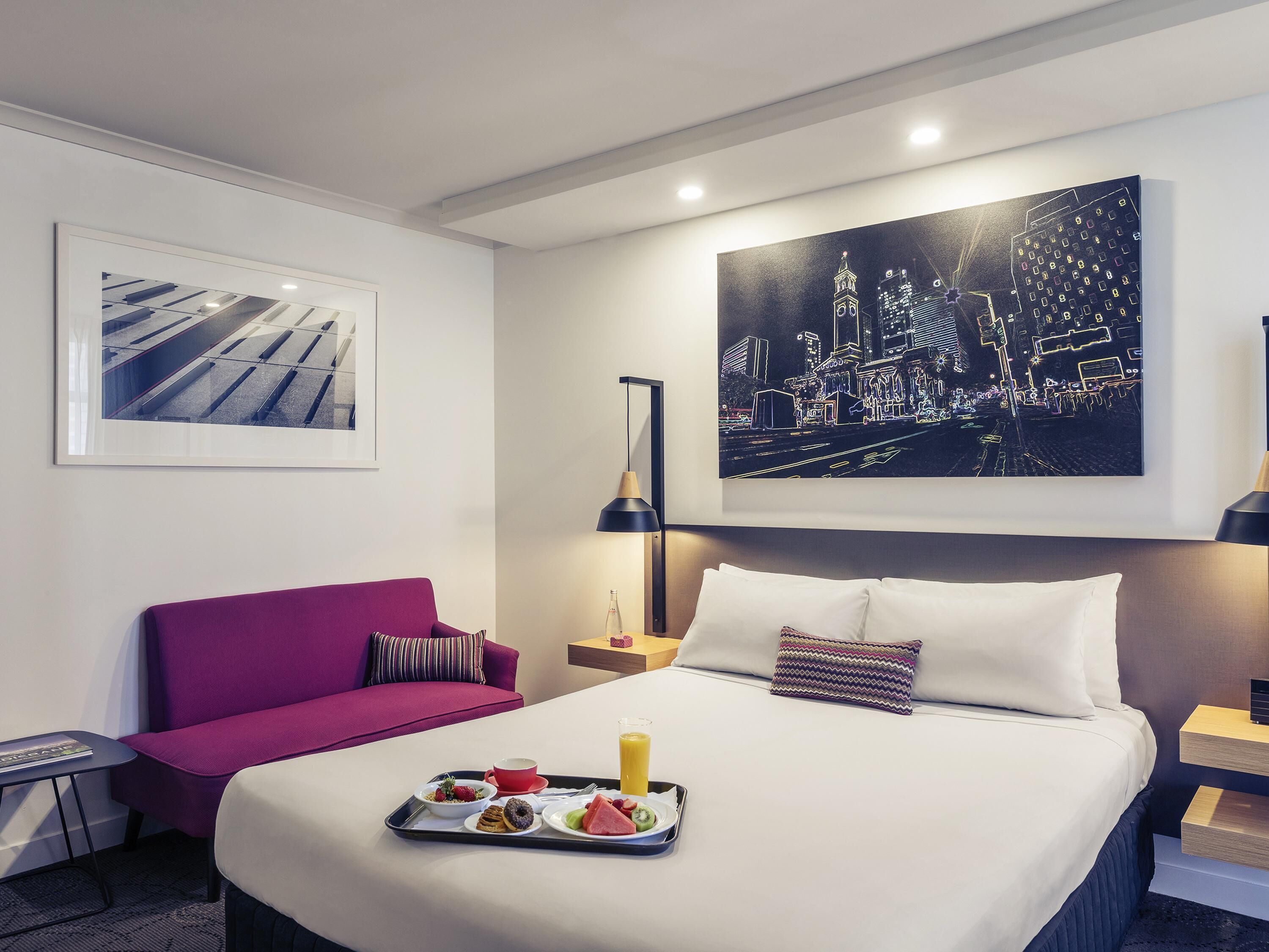 A hotel room with a bed, a purple sofa, a breakfast tray, and two artworks on the walls.