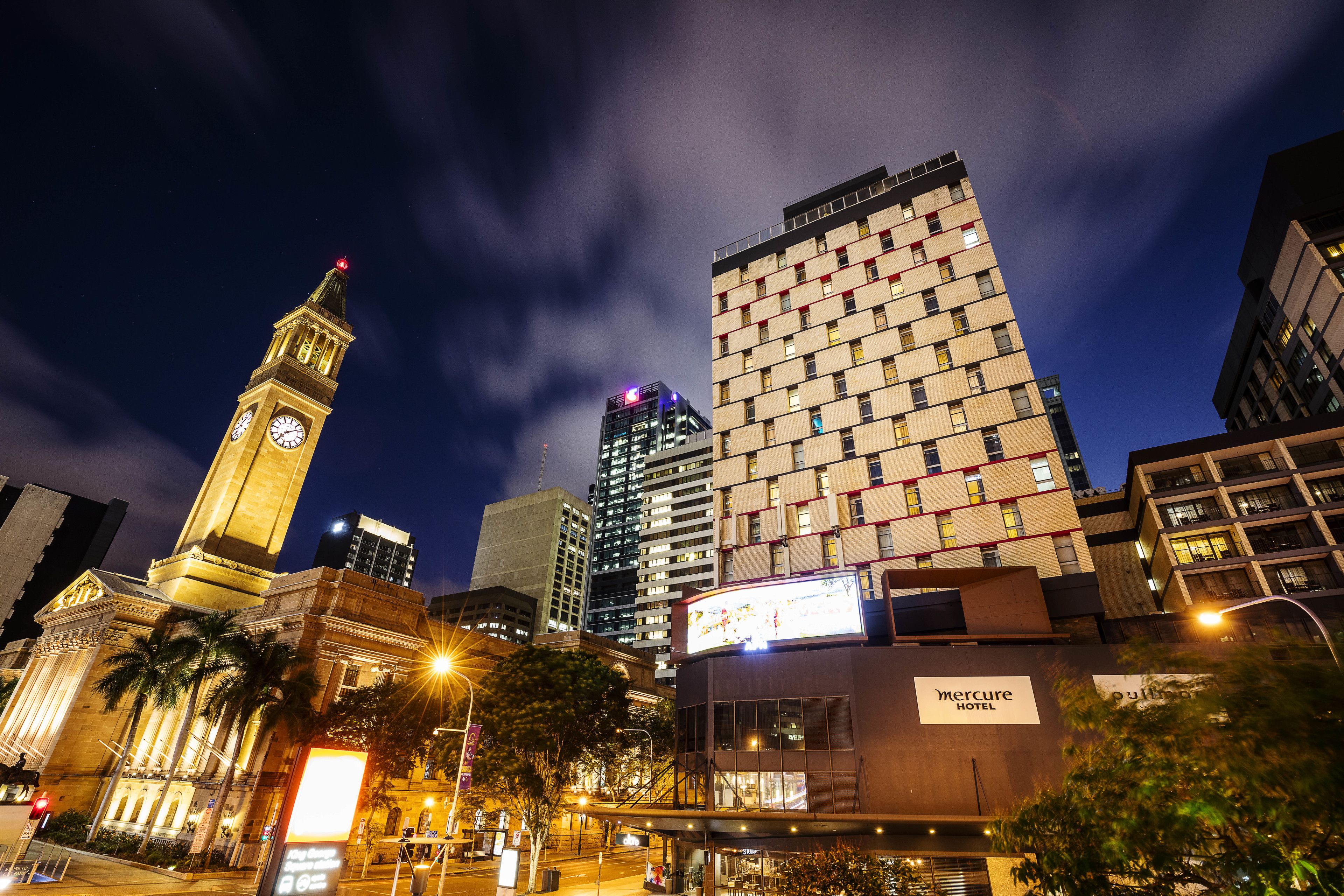 A modern hotel with a prominent clock tower in the background, surrounded by high-rise buildings.
