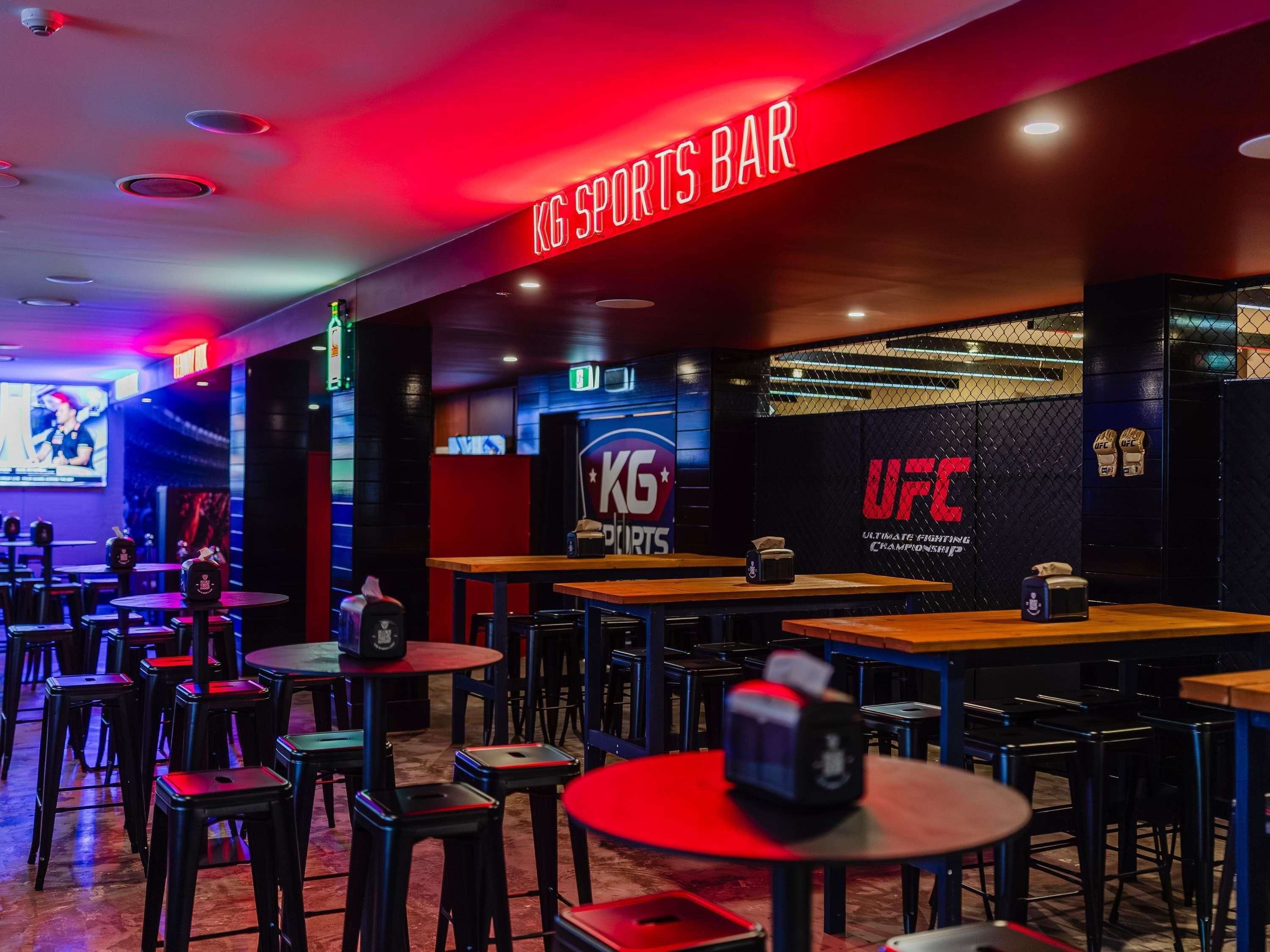A sports bar with multiple tables and stools, a large screen displaying sports content, and a prominent UFC logo on the wall.