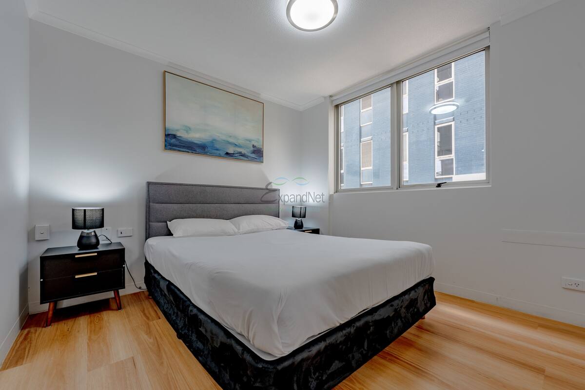 Ein Schlafzimmer mit Bett, Nachttisch, Lampe, einem Gemälde und einem Fenster mit Jalousien.