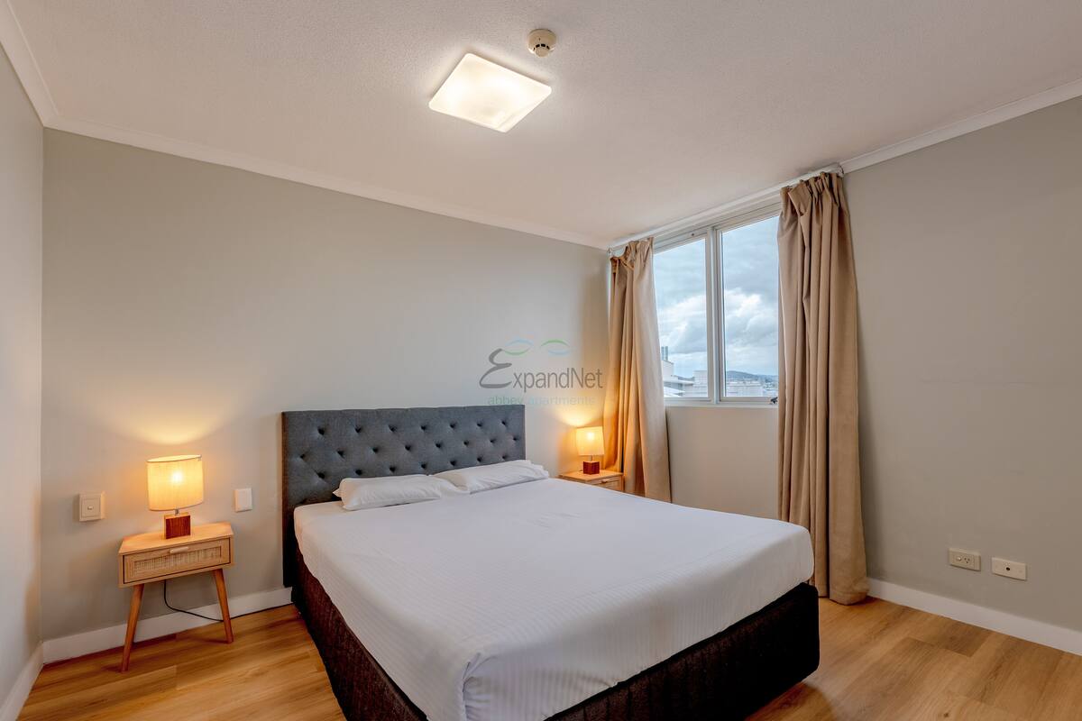 Ein Hotelzimmer mit einem großen Bett, zwei Nachttischlampen, einem mit Vorhängen versehenen Fenster und einer Deckenleuchte.