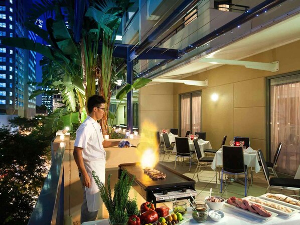 A chef cooking outdoors with a grill, surrounded by tables with chairs, and a cityscape in the background.