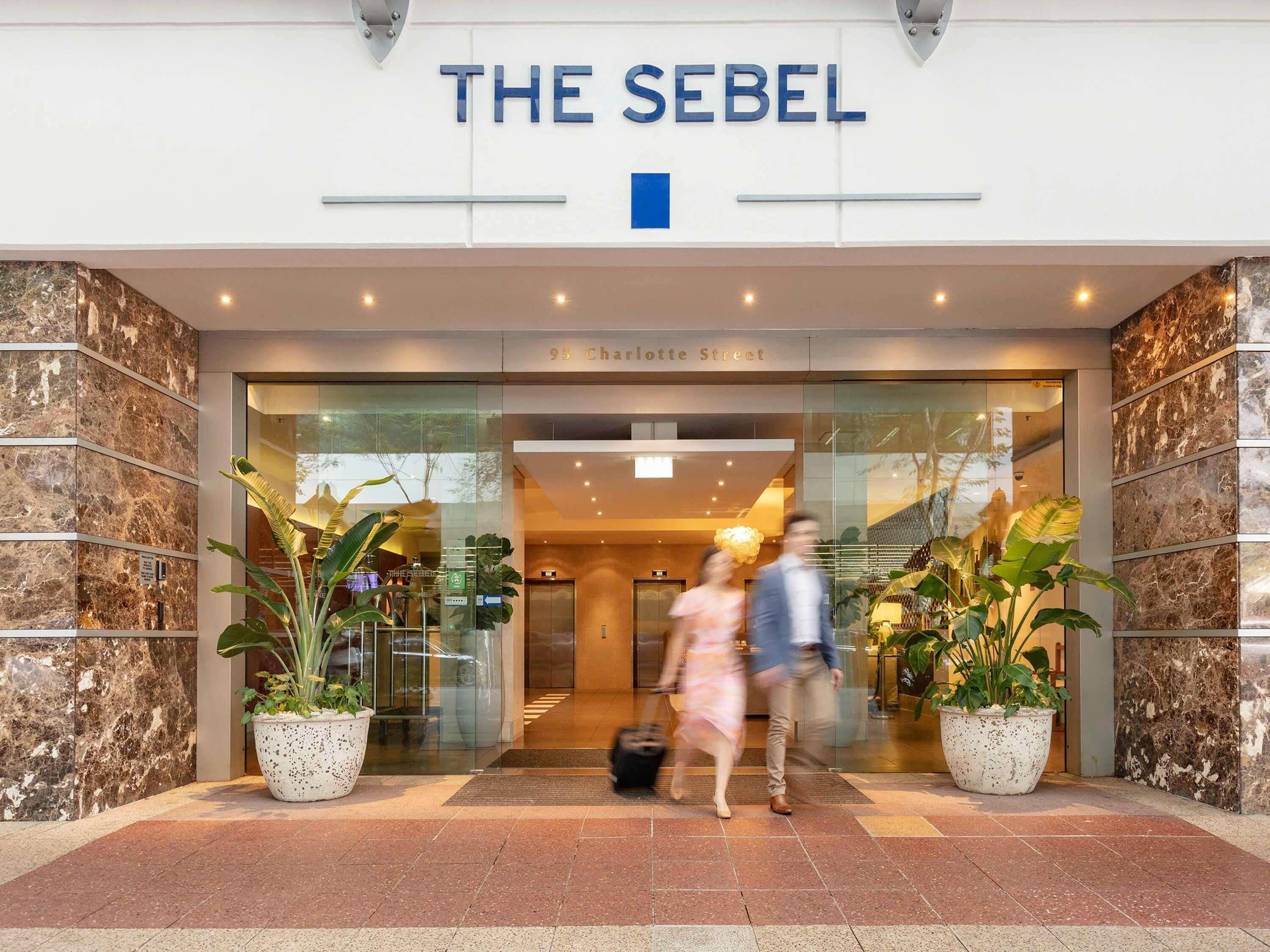 La entrada del hotel The Sebel, con fachada de mármol y plantas en macetas.