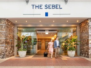 The entrance of The Sebel hotel with a marble facade and potted plants.