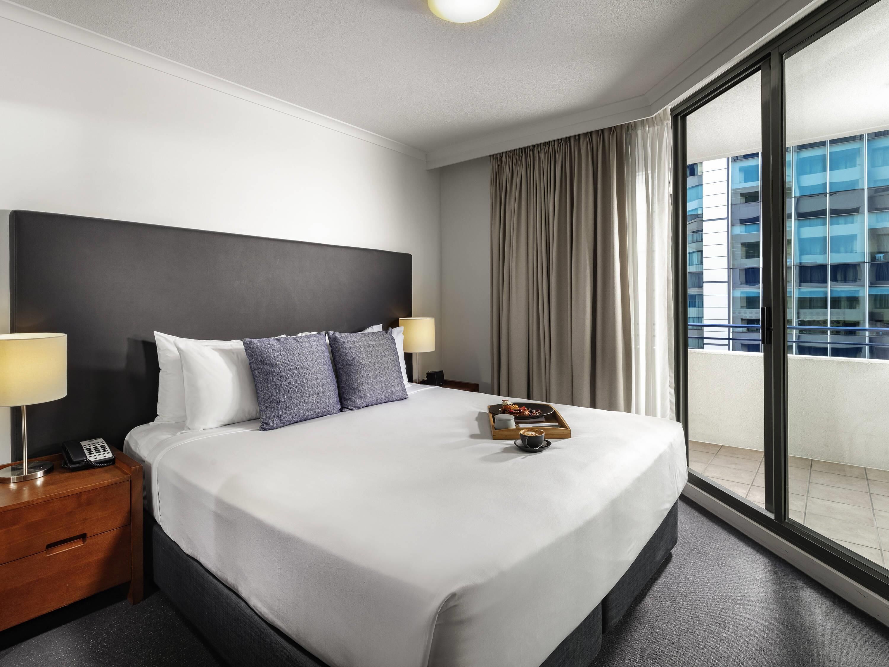 Ett hotellrum med en stor säng, två sängbord, en sänglampa, ett bricka med mat och en kopp, och utsikt över en byggnad genom ett stort fönster.