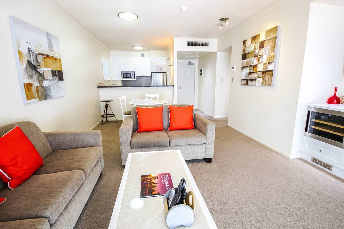 A modern living room with a grey sofa and a coffee table, a dining area with a white table and chairs, and a kitchen in the background.
