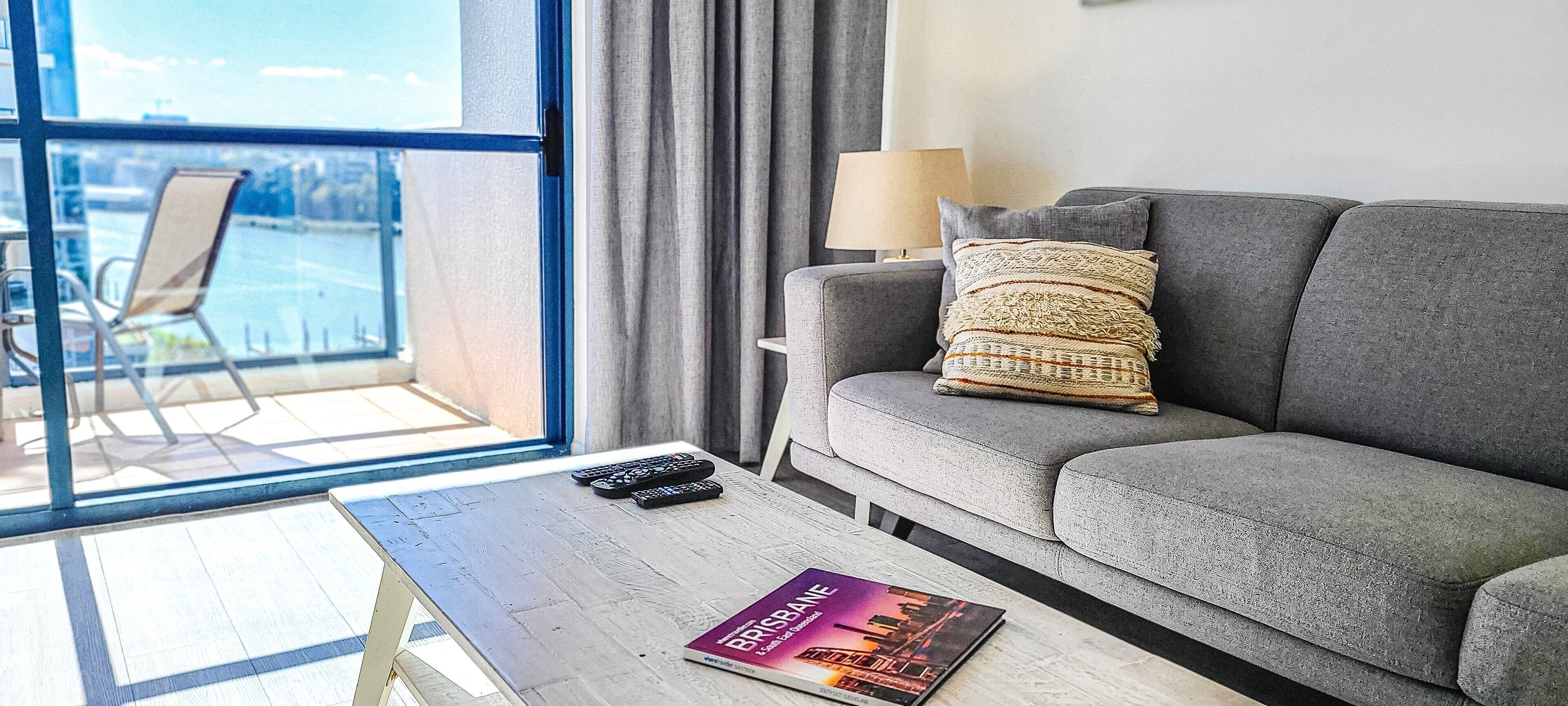 A modern living room with a grey sofa, a small table with a book and remotes, a lamp, and a view of a body of water through large windows.
