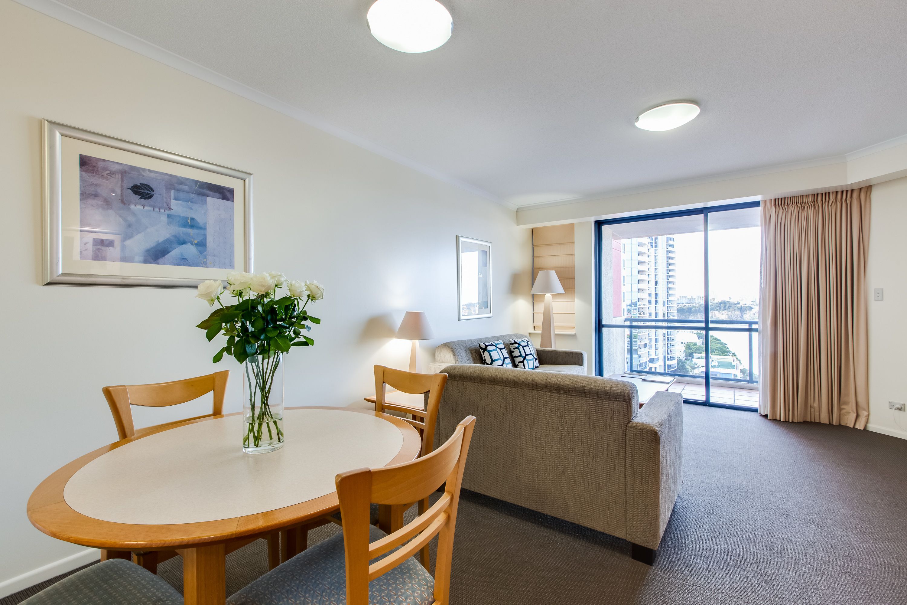 Habitación de hotel con una mesa de comedor, sillas, un sofá, un ventanal con vistas a edificios y un cuadro en la pared.