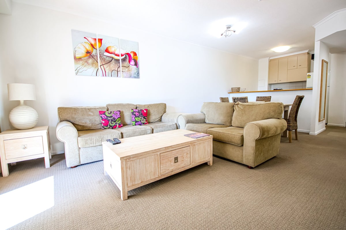 A living room with two sofas, a coffee table, and a lamp. There is a kitchen in the background.