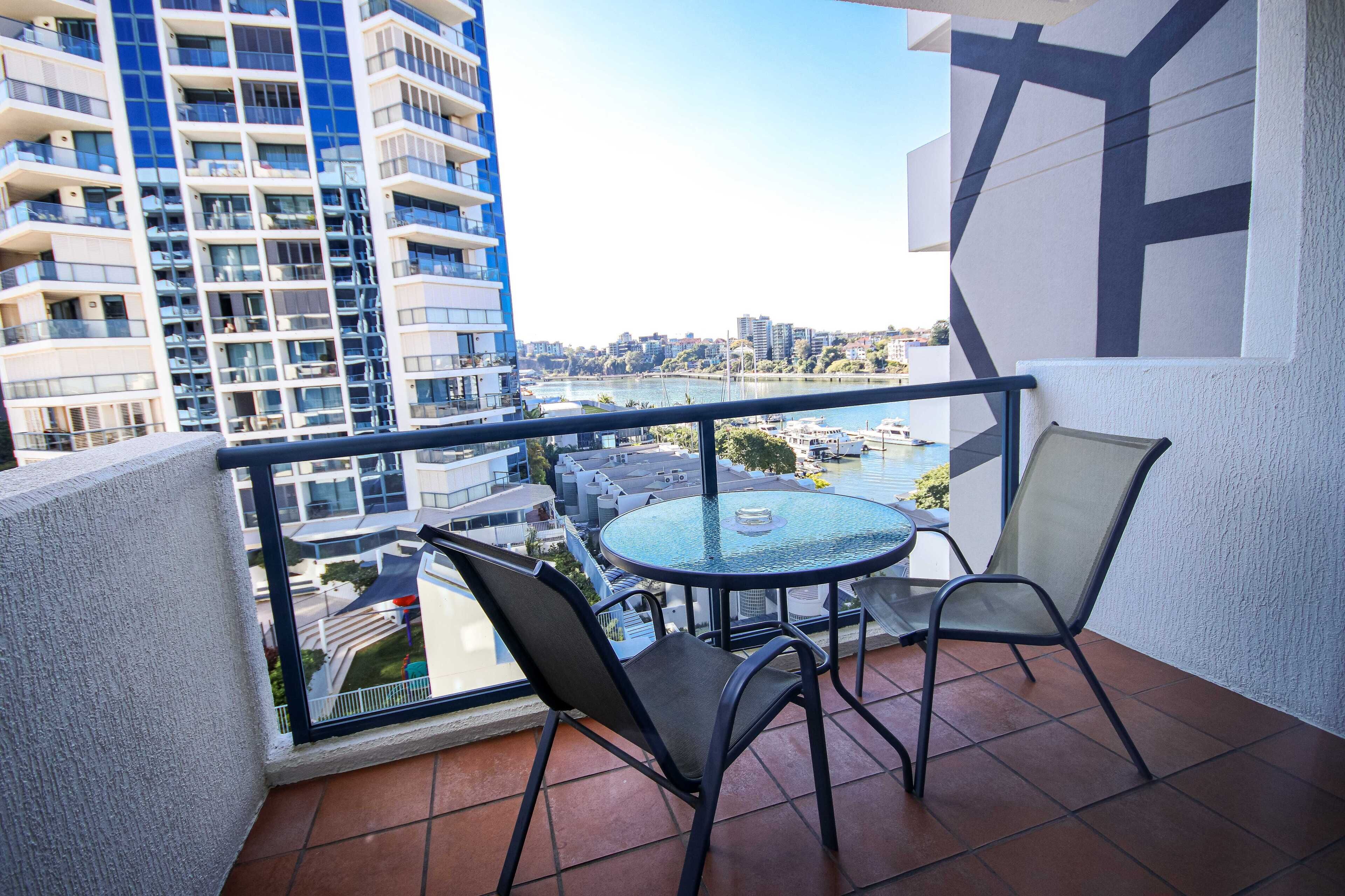 Un balcón con mesa y sillas de vidrio, con vistas a un río y al paisaje urbano.