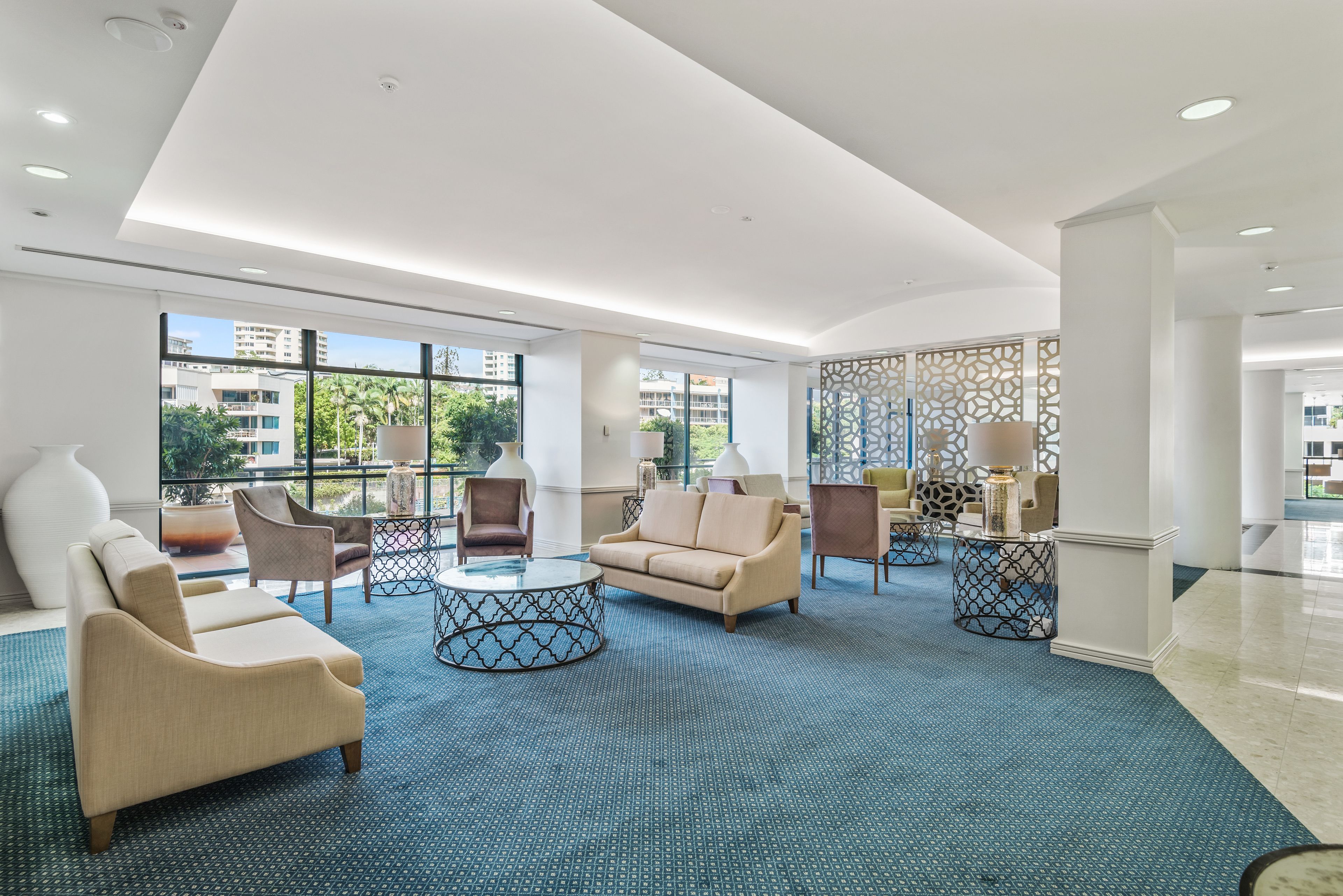A spacious lobby with a blue patterned carpet, beige sofas, and armchairs, large windows offering a view of buildings and greenery, and a decorative partition with a geometric pattern.