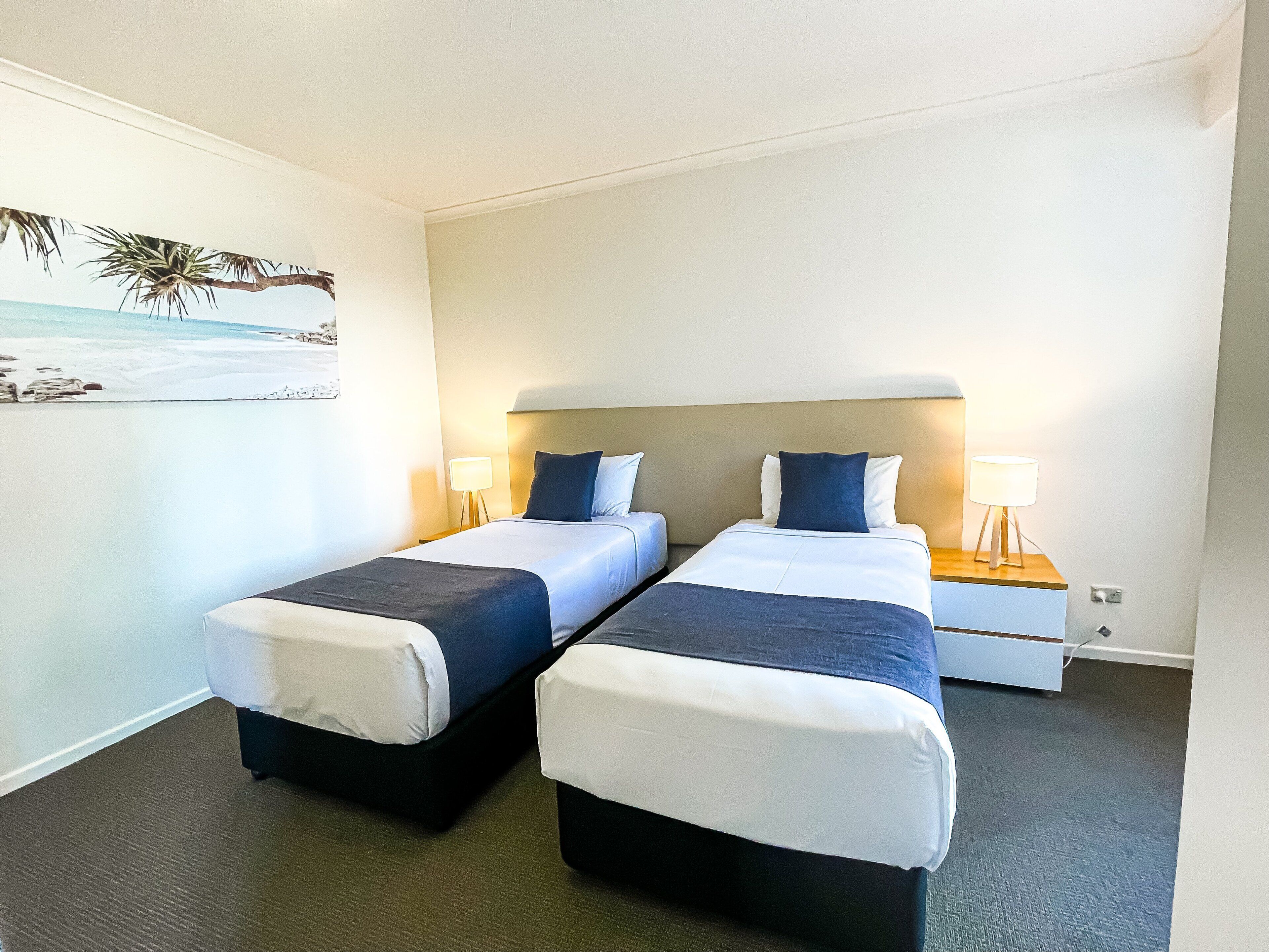 A hotel room with two beds, a headboard, bedside lamps, a painting of a beach scene, and a carpeted floor.