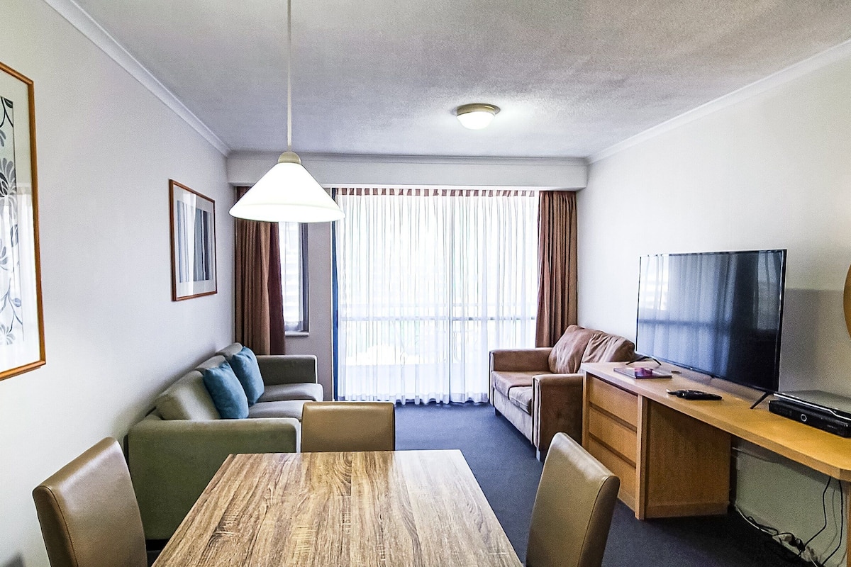A hotel room with a dining table, sofa, TV, and a large window with curtains.