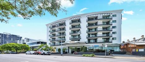 A multi-story hotel with a modern design, featuring balconies on each floor, a prominent entrance, and a parking area in front.