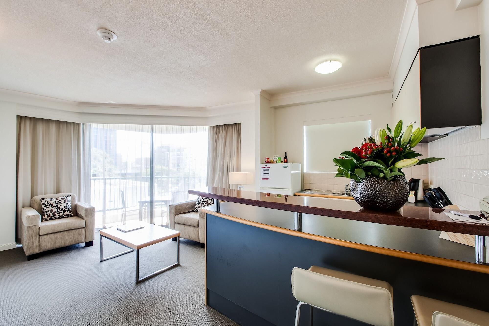 A hotel room with a living area, a kitchenette, a dining area, and a large window with curtains.