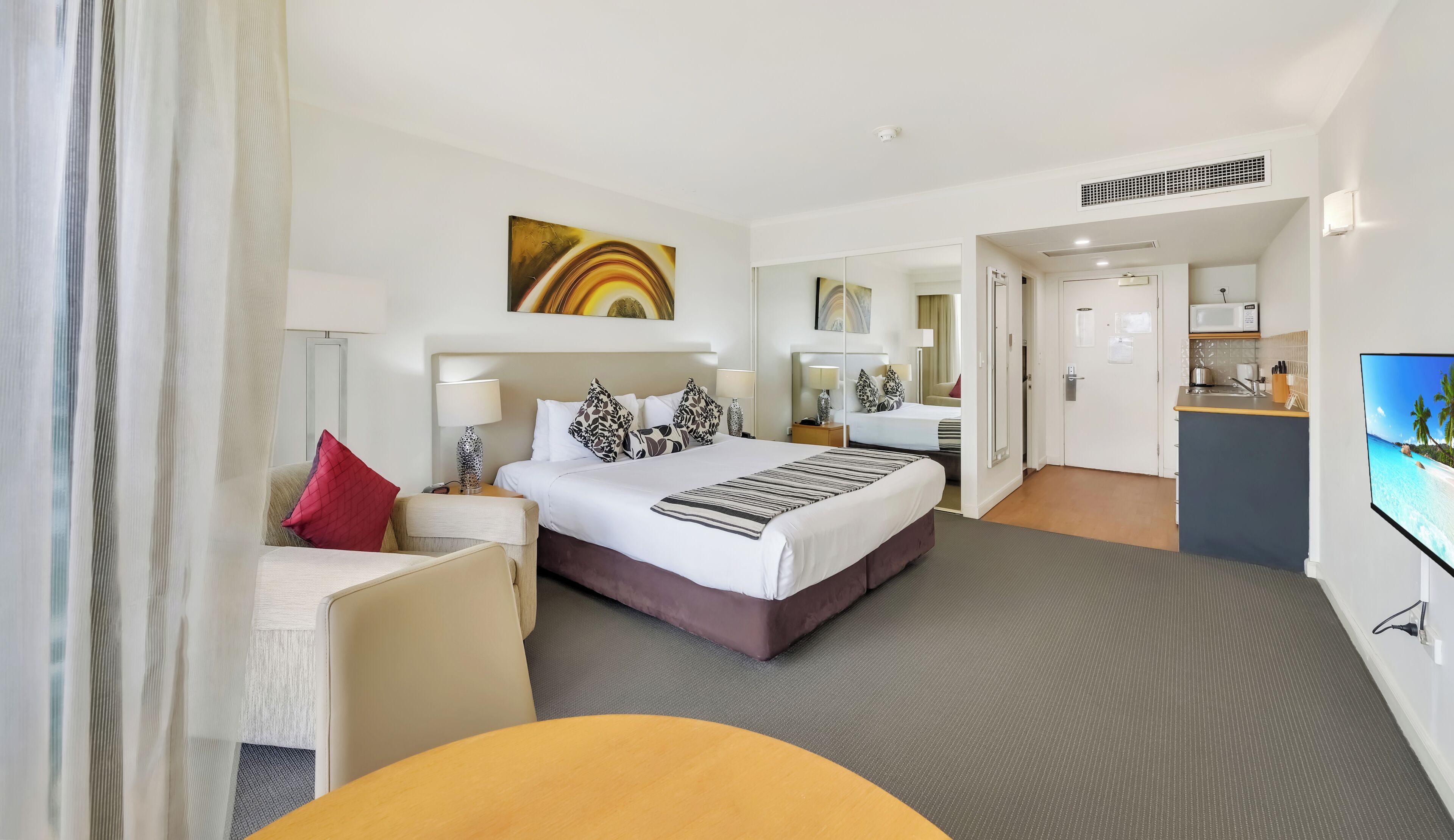 A hotel room with a bed, bedside tables, a TV, a desk, and a kitchenette.