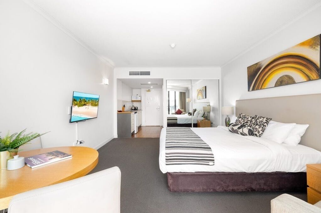 A hotel room with a bed, a desk, a TV, and a view of the kitchen and bathroom.
