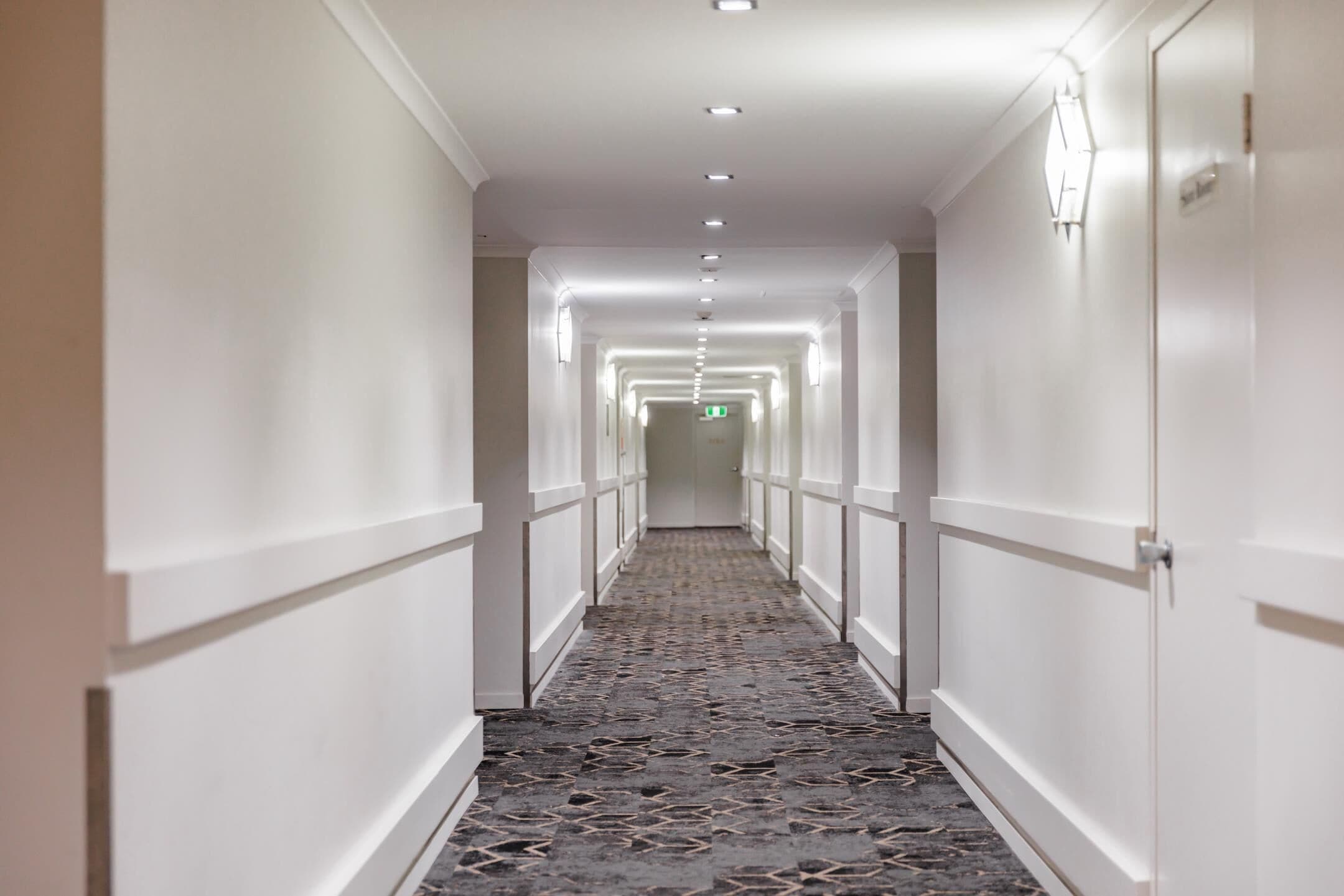 Un pasillo largo y estrecho en un hotel, con alfombra estampada, paredes blancas y puertas a ambos lados.