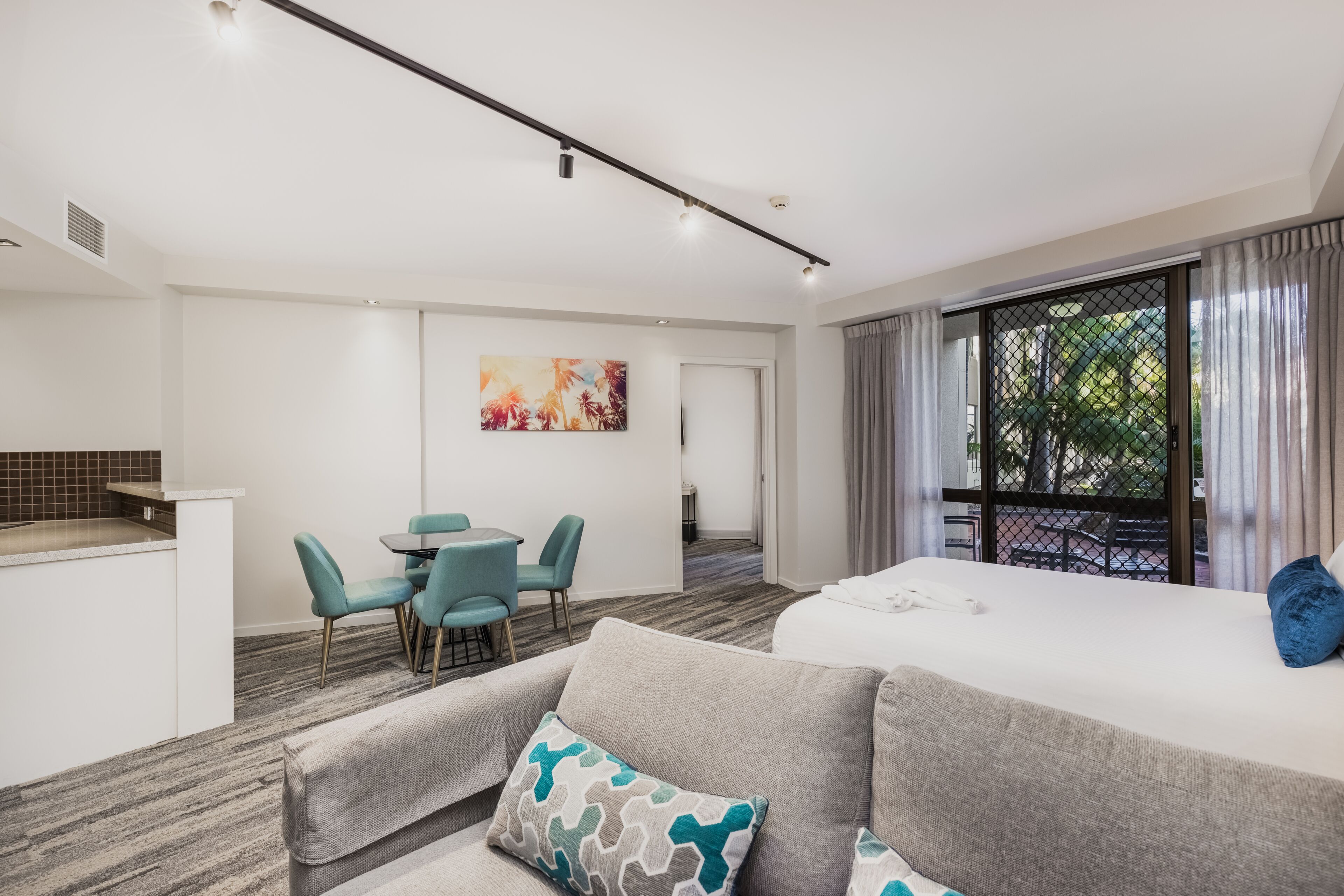 A modern hotel room with a sofa, a dining area with chairs, a kitchen area, and a large window with curtains.